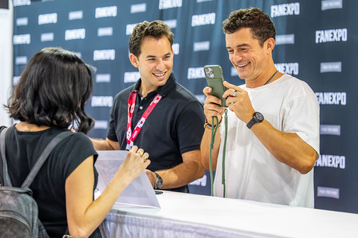 Orlando Bloom, wearing a casual white t-shirt, laughs as he takes a fan’s photo on his phone at the autograph table. A show staffer smiles beside him, while the fan watches from across the table, clearly delighted by the playful interaction.