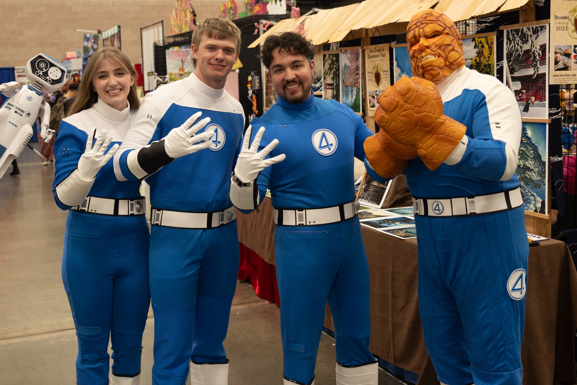 Four cosplayers dressed as the Fantastic Four stand together inside the convention hall, smiling and holding up four fingers, with one in a full Thing costume featuring orange rocky details.