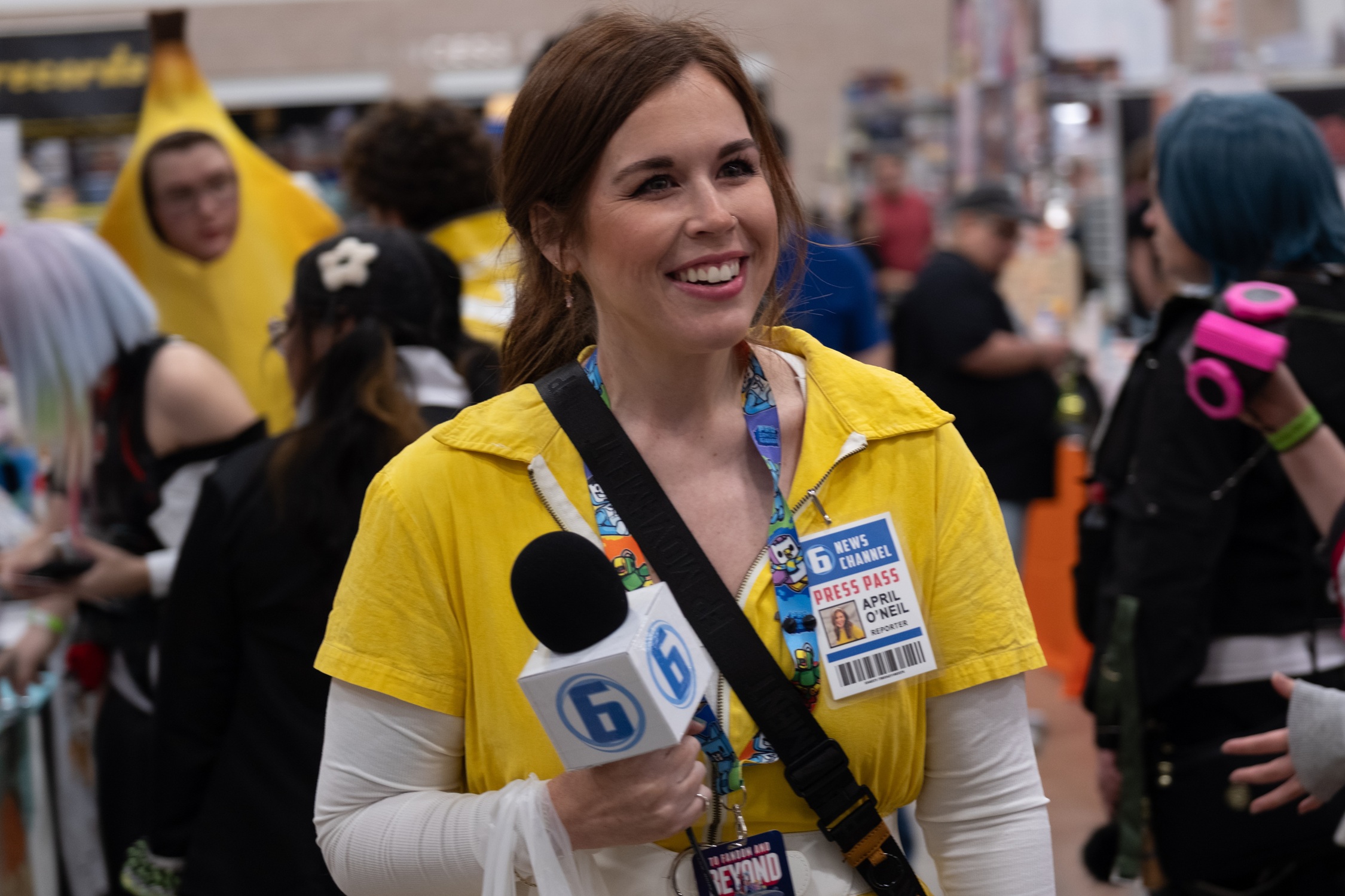 A cosplayer dressed as April O’Neil from Teenage Mutant Ninja Turtles smiles while holding a microphone and wearing a yellow jumpsuit with a press pass badge around her neck.