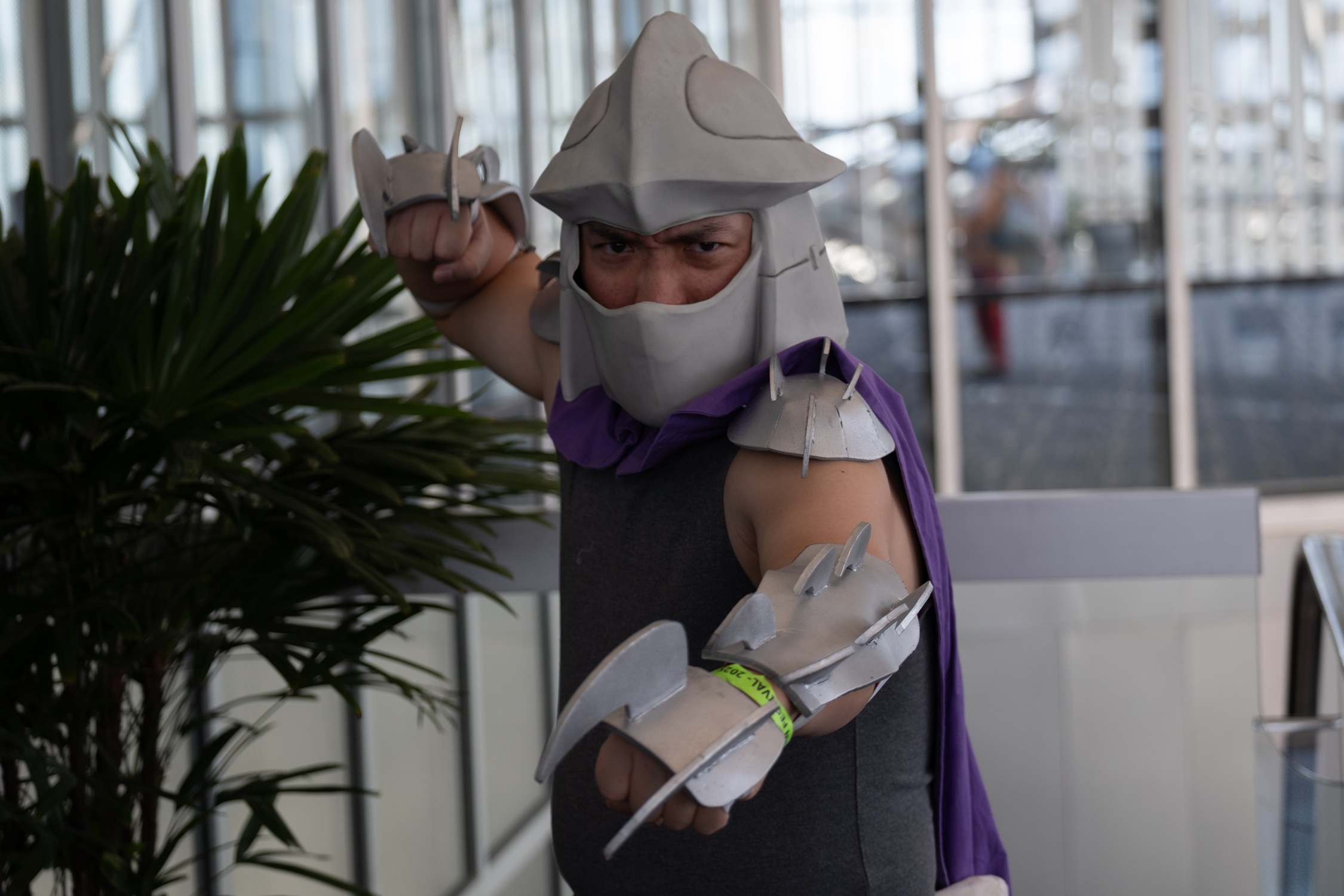 A cosplayer dressed as Shredder from Teenage Mutant Ninja Turtles poses dramatically with bladed gauntlets extended and a purple cape flowing behind them.