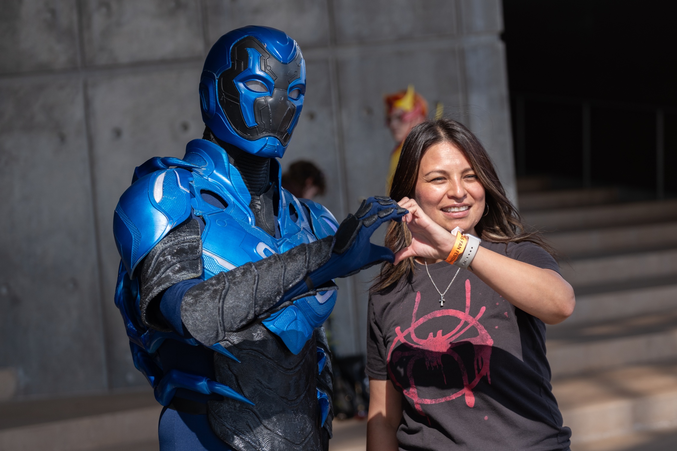 A cosplayer in a detailed Blue Beetle suit poses with a fan wearing a Spider-Verse T-shirt, both smiling and forming a heart shape with their hands.