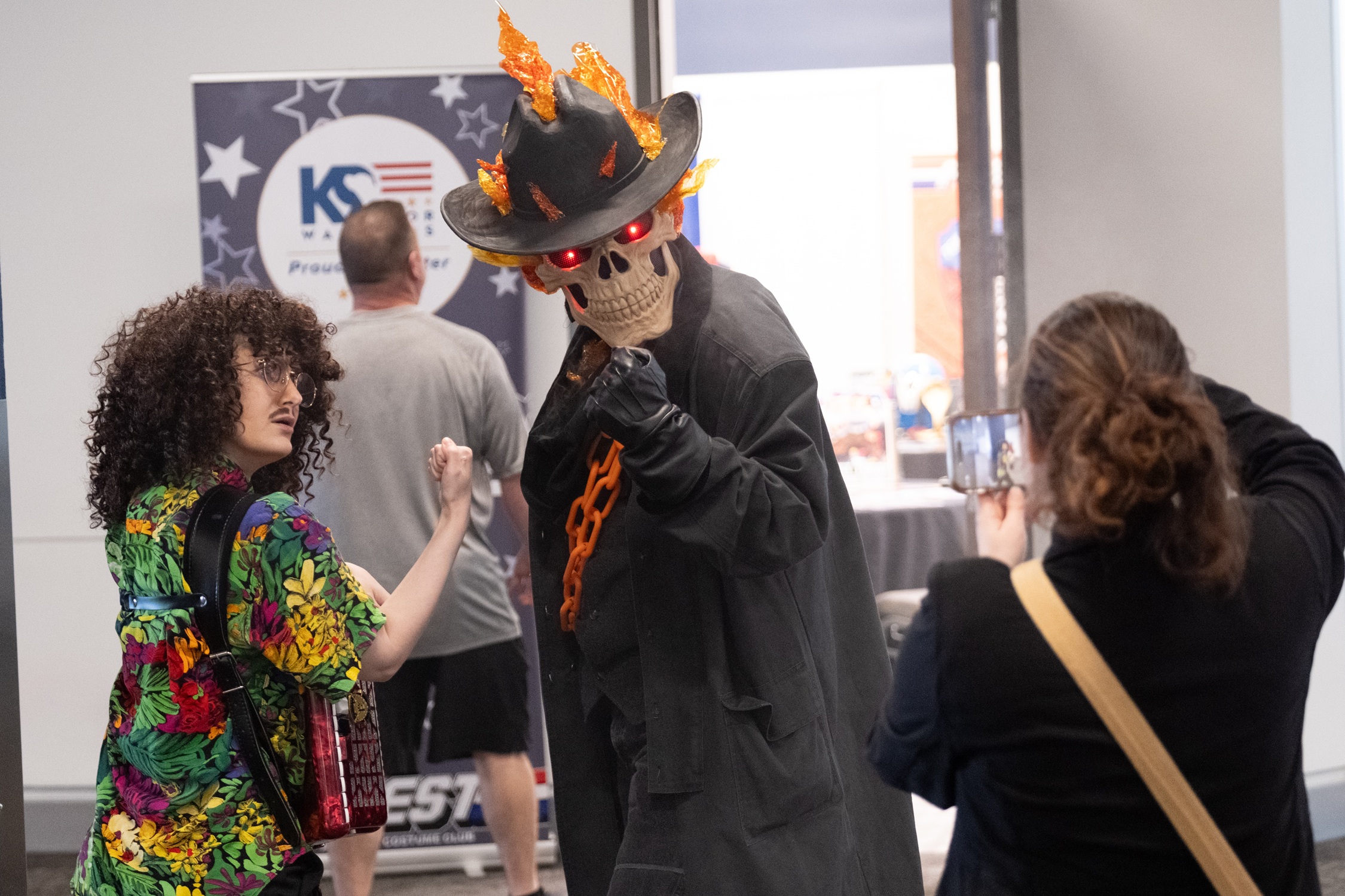 Two cosplayers interact on the show floor — one dressed as “Weird Al” Yankovic with curly hair and a colorful shirt, and the other as Ghost Rider with a flaming skull mask and glowing red eyes, while a bystander takes a photo.