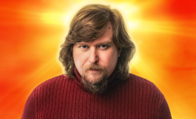 A man with long. dirty blonde hair, and a wispy beard, wearing a red turtleneck sweater stands in front of a yellow and orange background that appears as though light is appearing from behind his head.