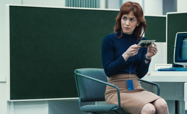 Helly sits in a chair at her cubicle. She holds a photo of four men, looking at someone off screen. The photo is turned toward the camera, indicating that she was inspecting the back side.