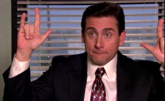 Michael Scott sits in his office, in front of the window. He does the 'rock on' gesture with his hands.