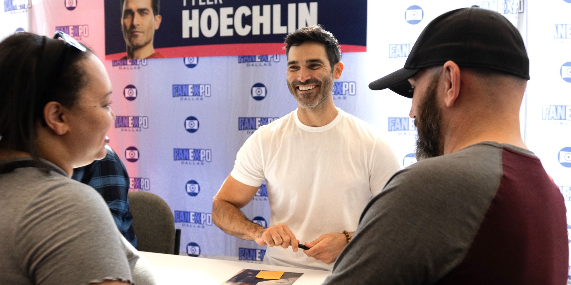 Actor Tyler Hoechlin smiles warmly while signing an autograph for fans at FAN EXPO Dallas. He sits at a table in front of a branded step-and-repeat backdrop, chatting with attendees as they present items for him to sign.