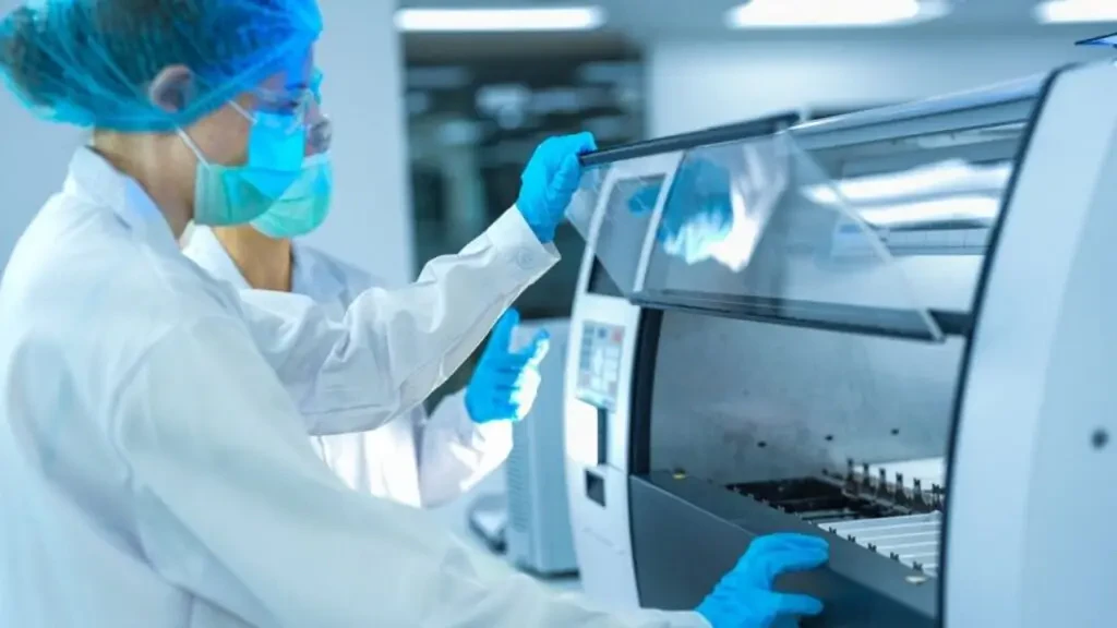 Profissionais de laboratório usando jaleco, máscara e luvas operando equipamento moderno de automação para diagnósticos clínicos e análises de saúde.