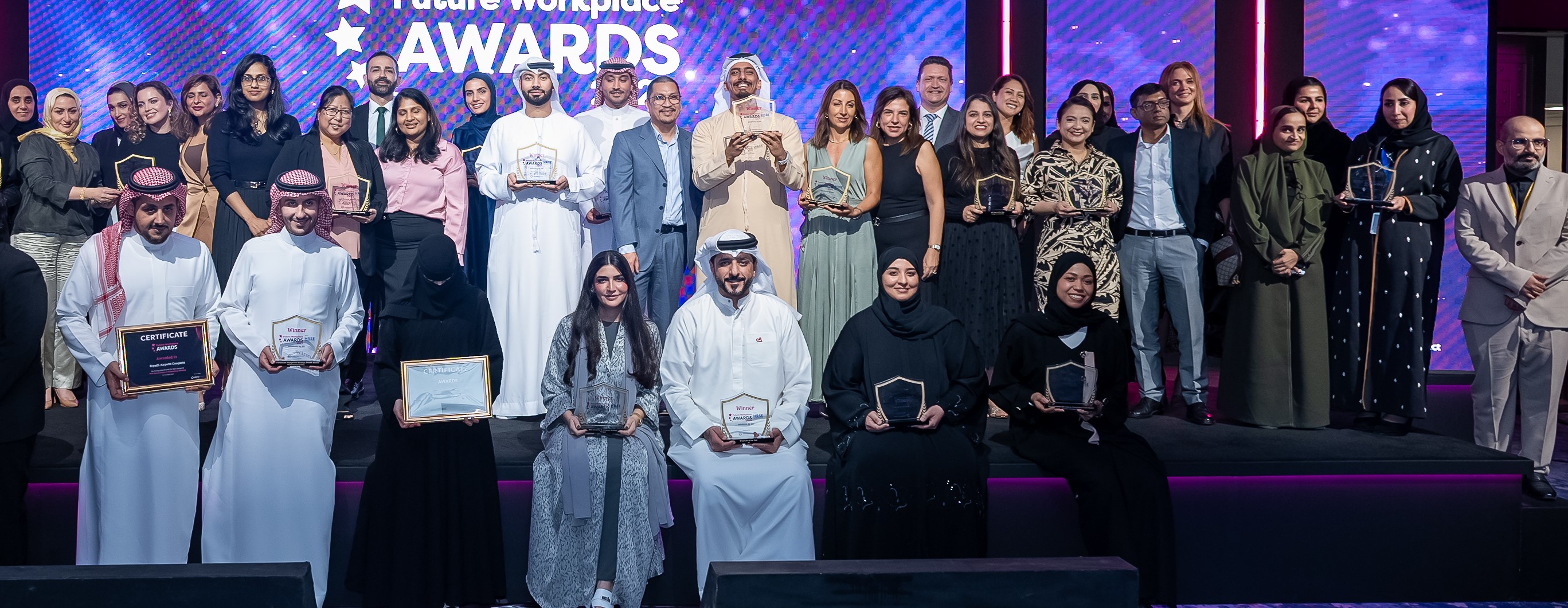 Conference attendees seated at round tables in a large meeting room, listening attentively, with notebooks and water glasses on tables.