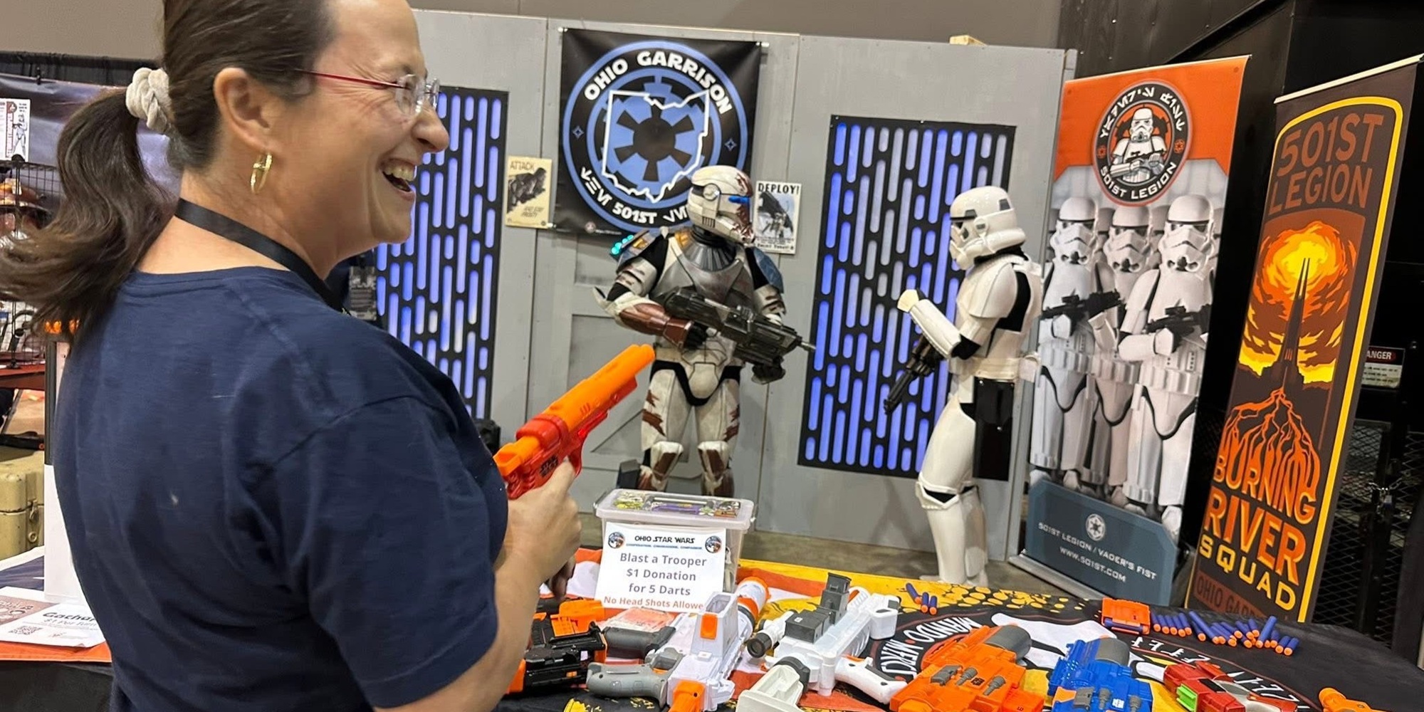 A smiling FAN EXPO Cleveland attendee aims a bright orange Nerf blaster toward costumed Star Wars troopers at the Ohio Garrison 501st Legion booth, with themed banners, props, and blasters displayed on a table in front of her.
