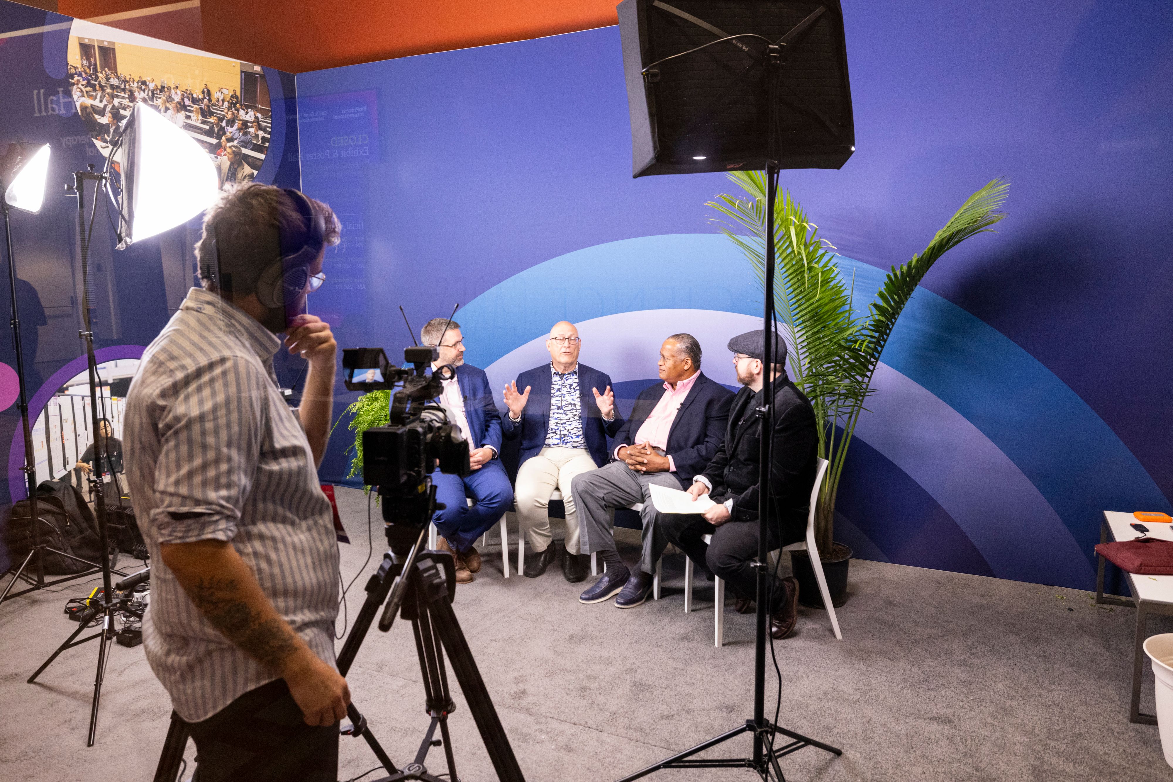 In a studio setup, four male speakers sit on chairs in discussion as a videographer films them at BioProcess International. There are large studio lights and a blue background with a palm plant next to the speakers.