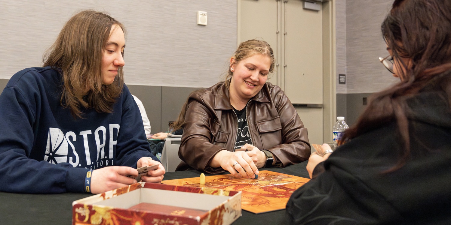 Three attendees sit at a table playing a board game, smiling and interacting as cards and pieces are spread between them. Shelves and stacks of games appear nearby in the board game library area.