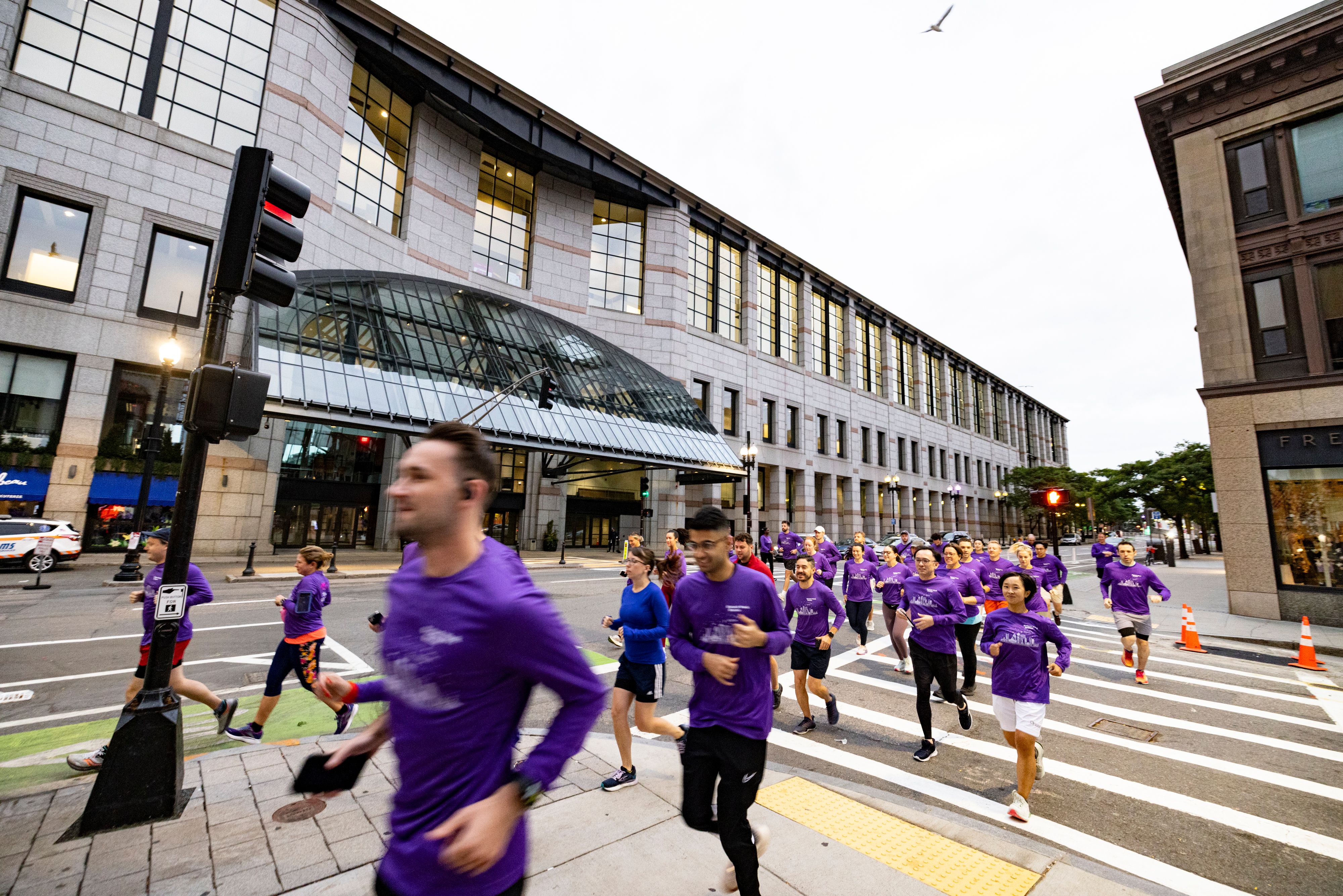 Bioprocessing and therapeutics professionals wearing matching purple t-shirts take part in a Fun Run, jogging across a road in Boston for BioTech Week Boston.