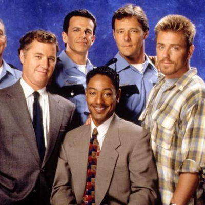 Giancarlo in a brown suit and multi-color checkered tie sitting for a picture with four men standing around him (2 wearing blue police uniforms, one in a gray suit and the other in a yellow/gray flannel)