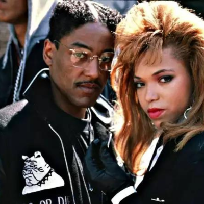 Giancarlo in a black bomber jacket with a bulldog on it. He is standing next to a girl with teased hair and they are both looking intensely at the camera.