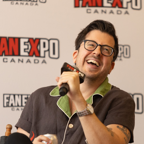 Christopher Mintz-Plasse smiles and laughs as he answers fan questions. He is sitting in front of a white background with "FAN EXPO Canada" logos all over it. He is holding a black FAN EXPO Canada branded microphone. 