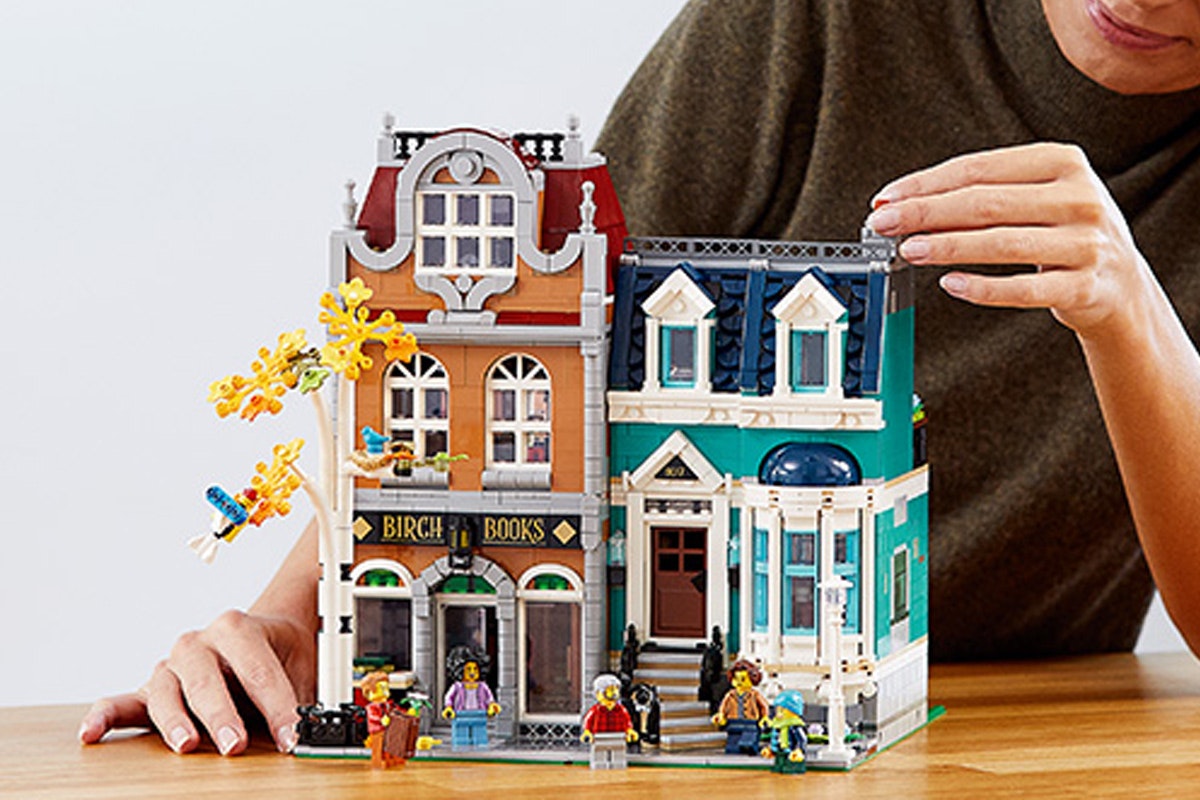 Two Lego buildings. One is an orange brown one with a sign that says Birch Books. Next to it is a teal building. There is a side walk with a birch tree with orange leaves and next to it are people walking around.