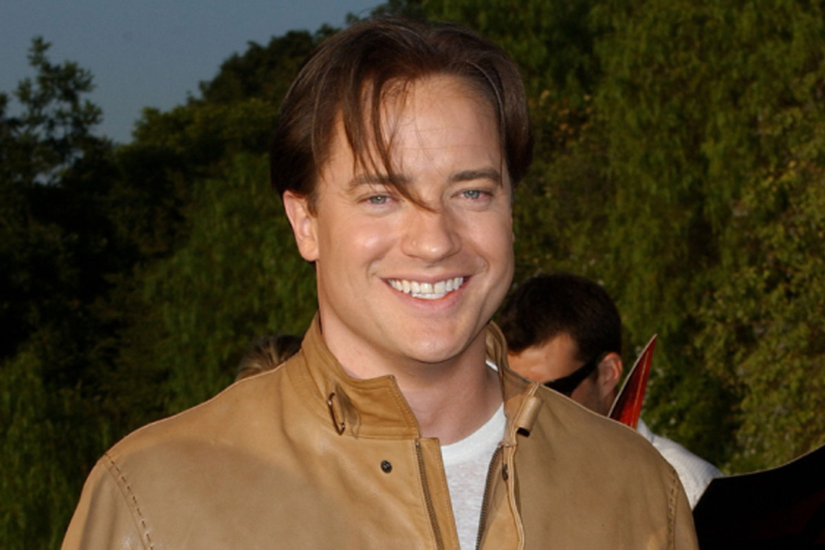 A younger Brendan Fraser smiles at the camera near some hedges while wearing a brown leather jacket