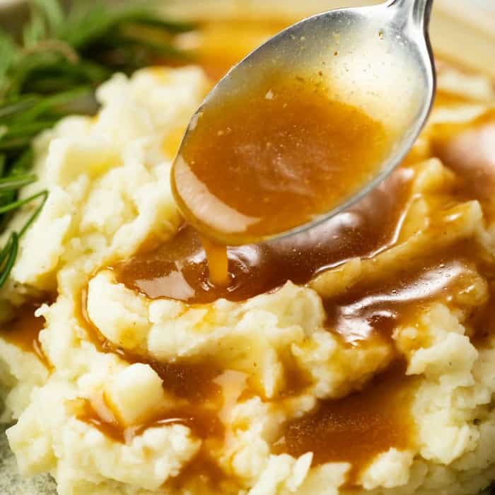 A silver spoon pours brown gravy onto a pile of white mashed potatoes