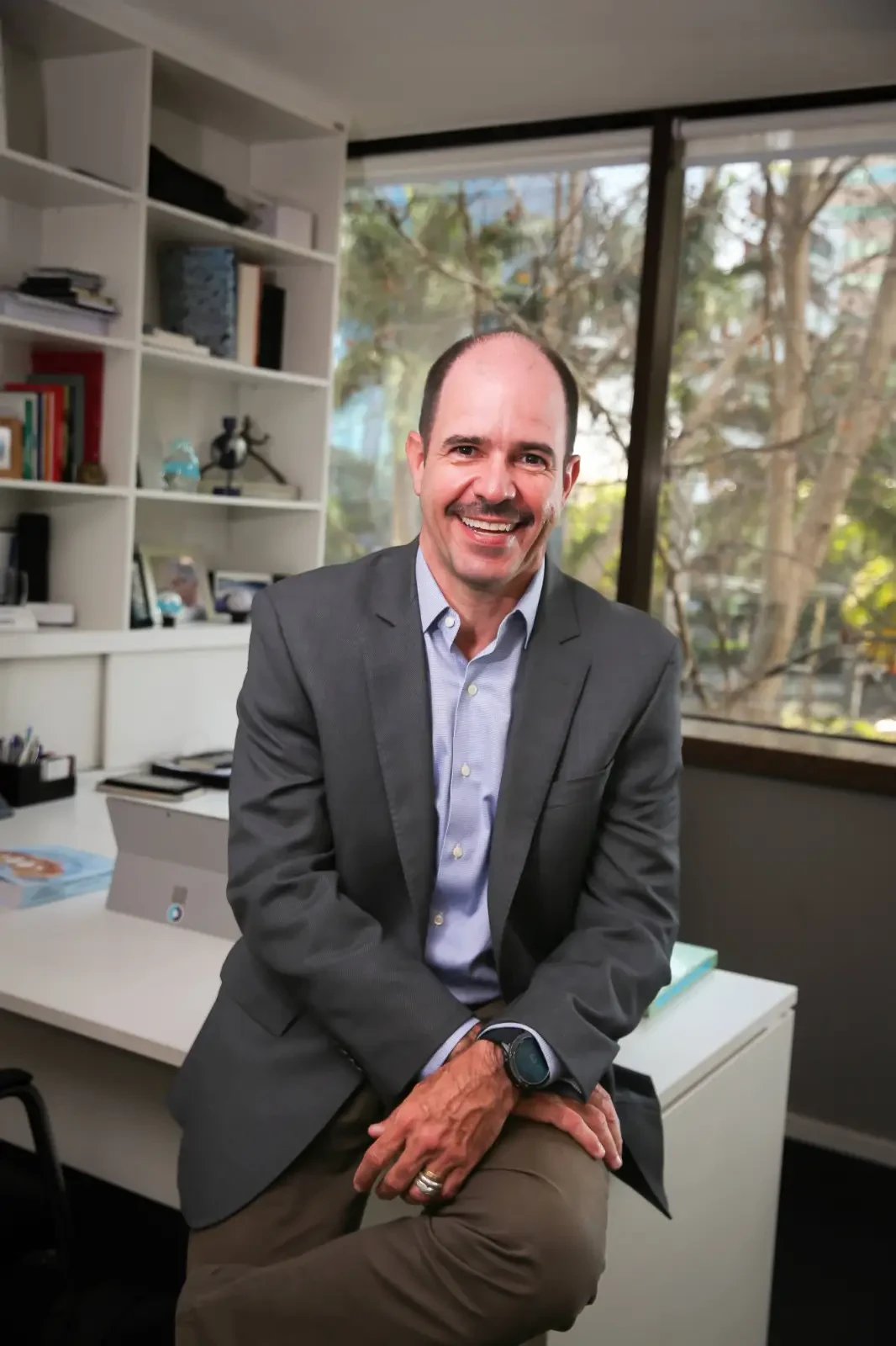Caio Soares, palestrante do Healthcare Innovation Show, homem sorrindo de bigode e blazer cinza em escritório.