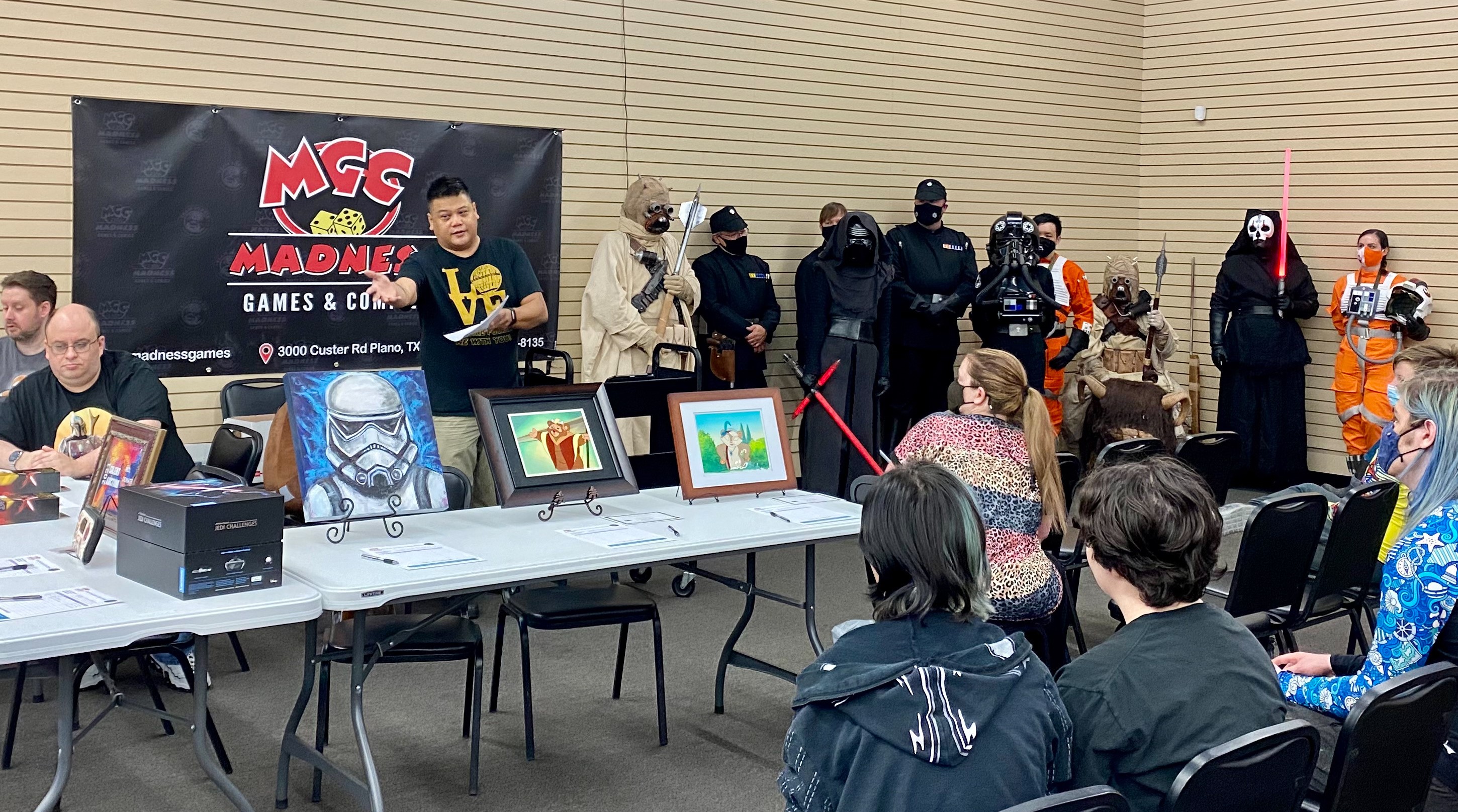 People looking at a variety of art work with Star Wars Cosplayers standing in the back. Man in middle in black shirt is gesturing to the left most artwork in front of him.