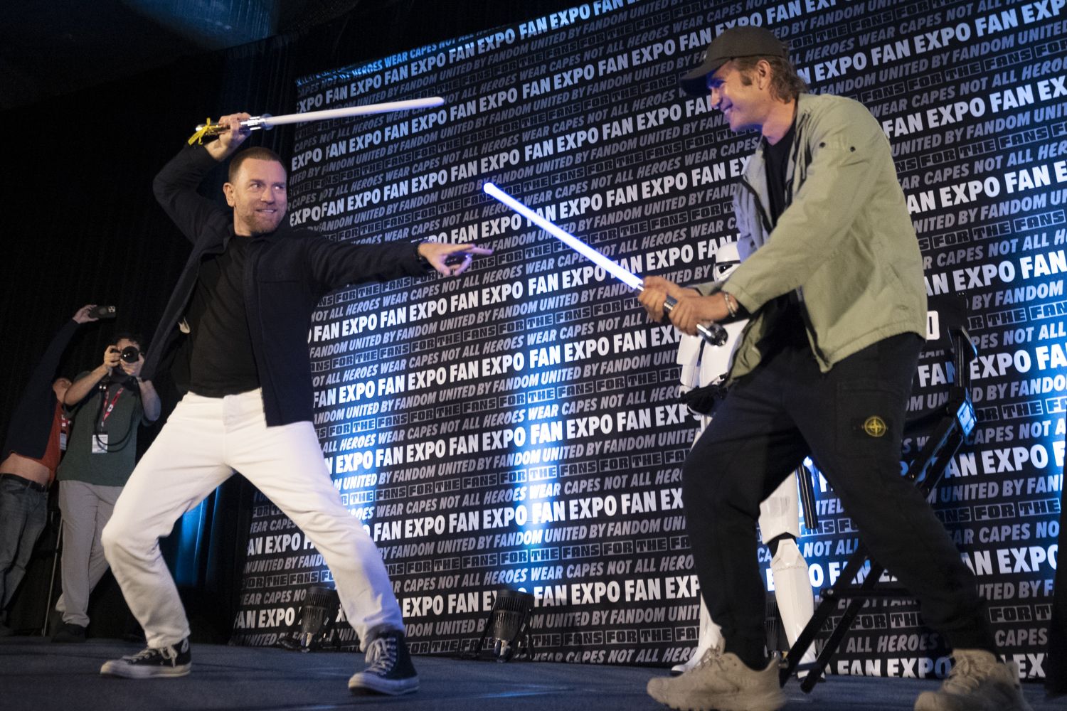 Ewan McGregor and Hayden Christensen face off on stage in a playful lightsaber duel, both grinning as fans cheer them on. A stormtrooper and Darth Vader stand in the background.