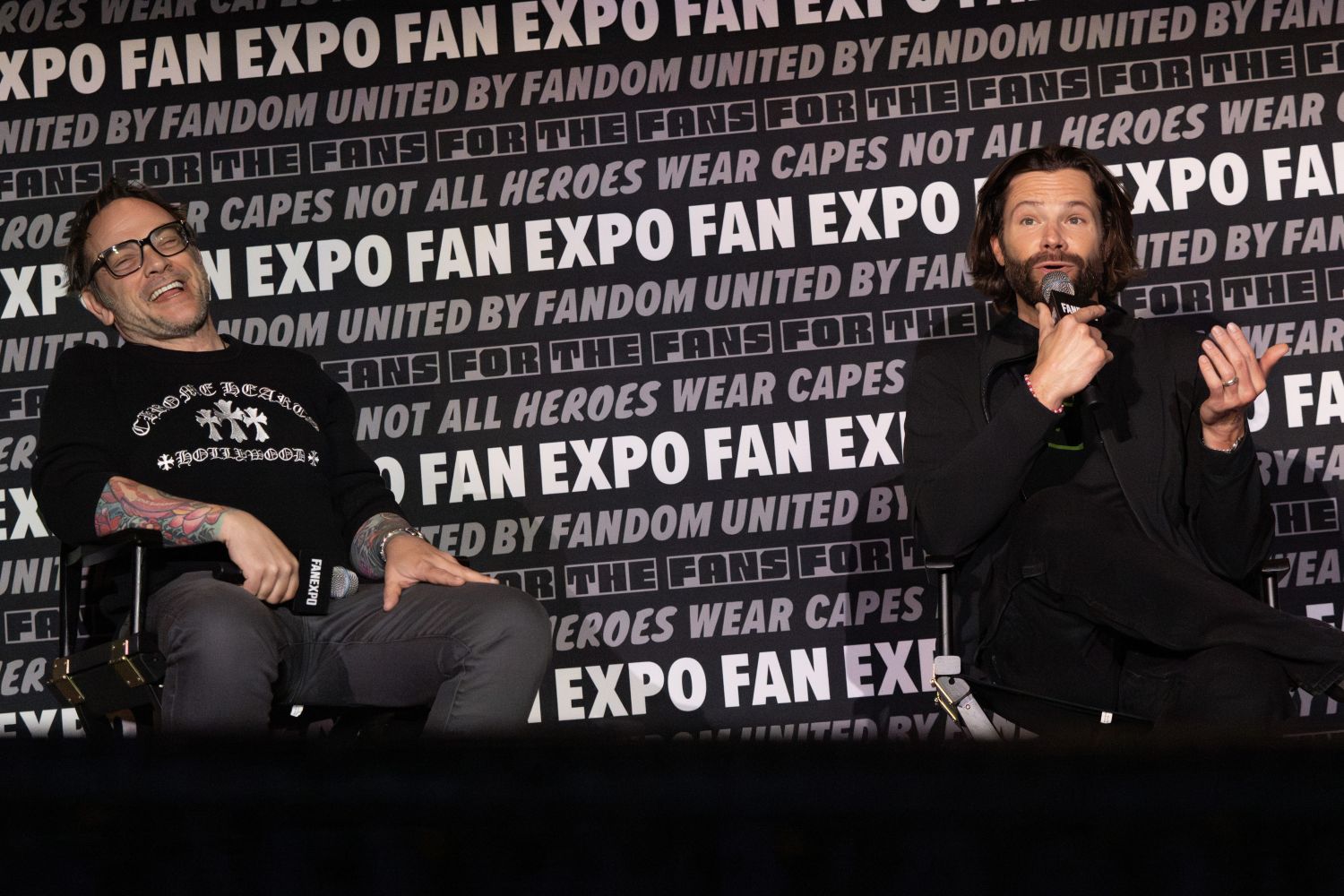 A humorous moment onstage between Mark Sheppard and Jared Padalecki. On the left, Mark Sheppard, with short hair, glasses, and colorful tattoos laughs while reclining in his chair, holding a FAN EXPO mic. He wears a black sweatshirt with white gothic lettering. On the right, Jared Padalecki gestures with one hand while holding a mic in the other, dressed in black with his signature long hair and beard. The FAN EXPO backdrop emphasizes fan unity and hero themes.