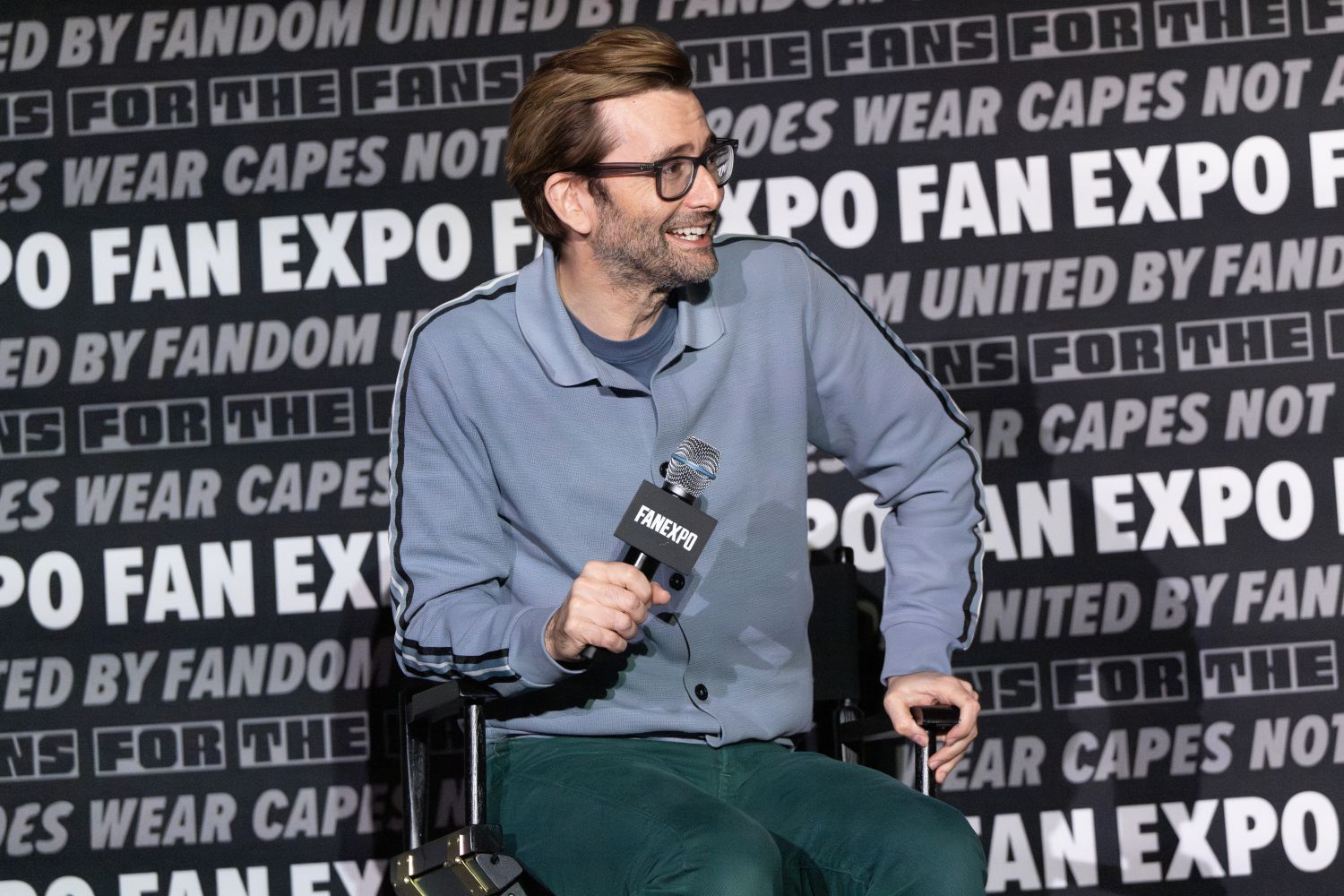 David Tennant, wearing glasses and a light blue zip-up jacket with black stripes, smiles as he looks to the side during a panel. He holds a microphone in one hand and leans forward slightly in his director’s chair. The wall behind him features FAN EXPO’s signature black-and-white branding with repeated phrases.