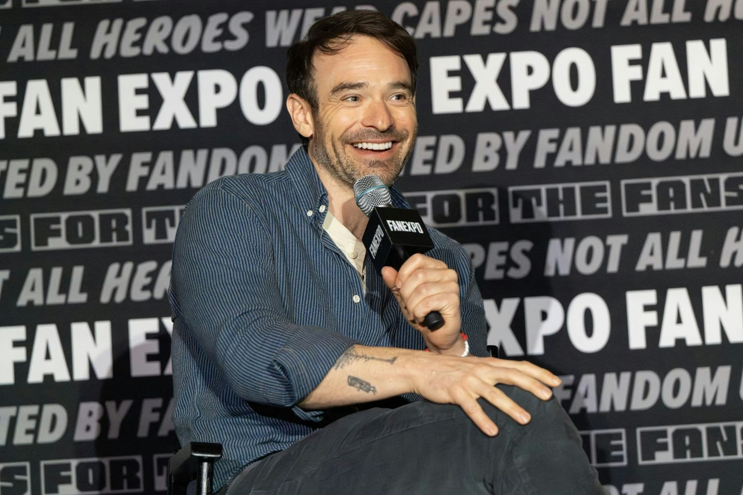 Charlie Cox sits onstage holding a microphone with a bright, cheerful smile. He wears a blue pinstriped button-up shirt with the sleeves rolled up and dark pants. A small tattoo is visible on his forearm. He gestures casually with one hand as he speaks. The background is filled with FAN EXPO logos and taglines.
