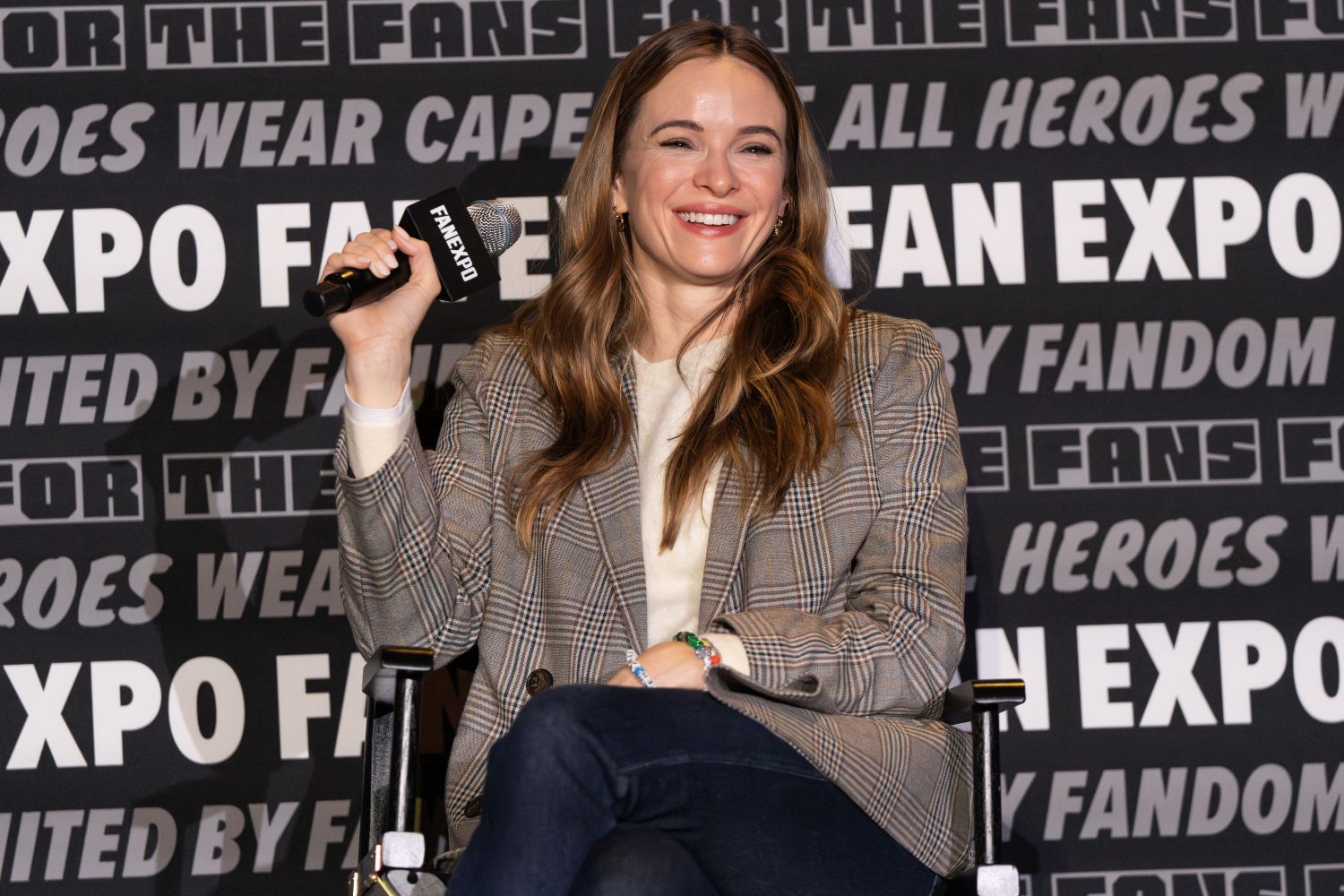Danielle Panabaker, with long, light brown hair sits on stage holding a FAN EXPO-branded microphone. She wears a beige sweater and a brown plaid blazer with dark jeans, seated in a director’s chair. Behind her, the black wall features bold white text repeating “FAN EXPO,” “FOR THE FANS,” and “UNITED BY FANDOM.”