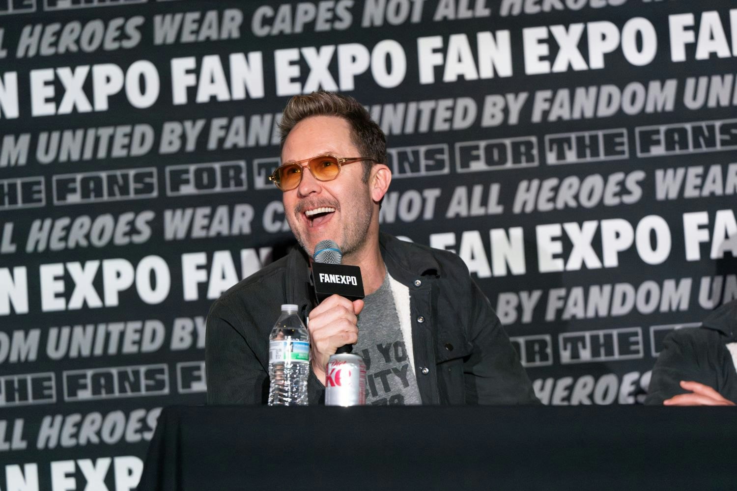 Michael Rosenbaum sits behind a panel table at FAN EXPO, mid-laugh as he holds a FAN EXPO-branded microphone in his right hand. He’s wearing amber-tinted glasses, a dark button-up shirt over a graphic tee, and appears relaxed and animated while speaking. In front of him on the table are a water bottle and a can of Diet Coke. The backdrop behind him features repeated phrases like “FAN EXPO,” “UNITED BY FANDOM,” and “NOT ALL HEROES WEAR CAPES” in bold, monochrome text.
