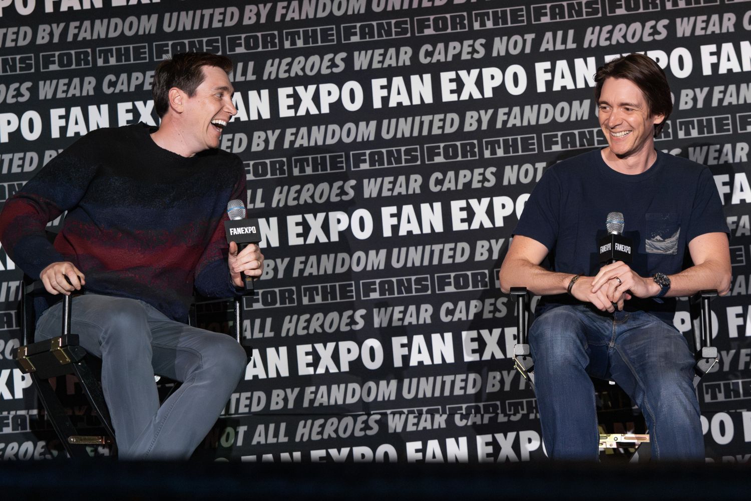 Twin actors James and Oliver Phelps sit in director’s chairs mid-conversation, laughing with microphones in hand. The man on the left wears a maroon and navy striped sweater and gray pants, leaning forward with a wide grin. The man on the right wears a navy t-shirt with a small chest graphic and jeans, smiling back. The FAN EXPO panel wall behind them is covered in repeated phrases about fandom and heroes.