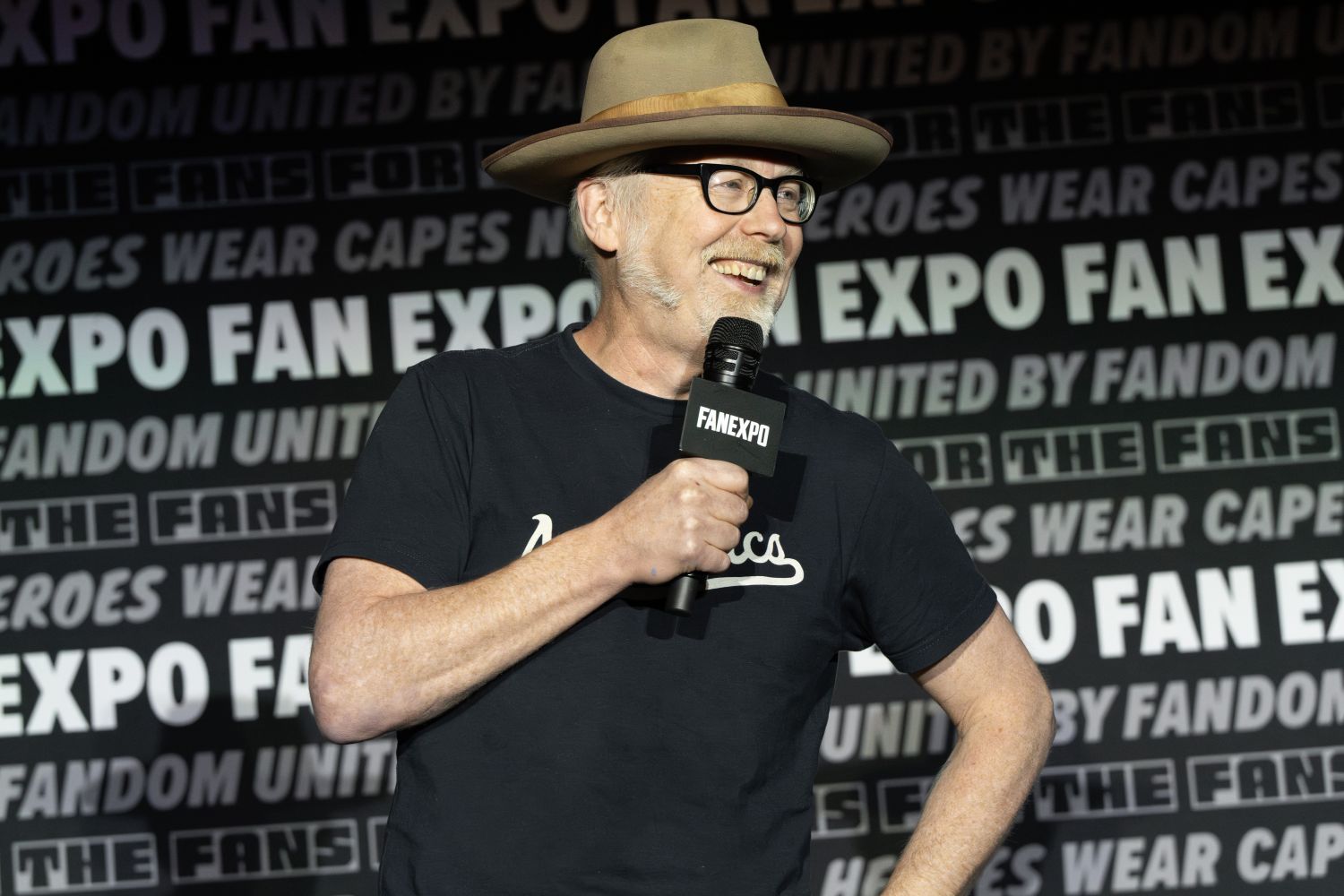 Adam Savage, with light skin, glasses, a trimmed white beard, and a tan wide-brimmed hat holds a microphone and smiles while speaking to the audience. He wears a black t-shirt with white script across the front. The FAN EXPO panel backdrop behind him includes phrases like “United by Fandom” and “Not All Heroes Wear Capes.”