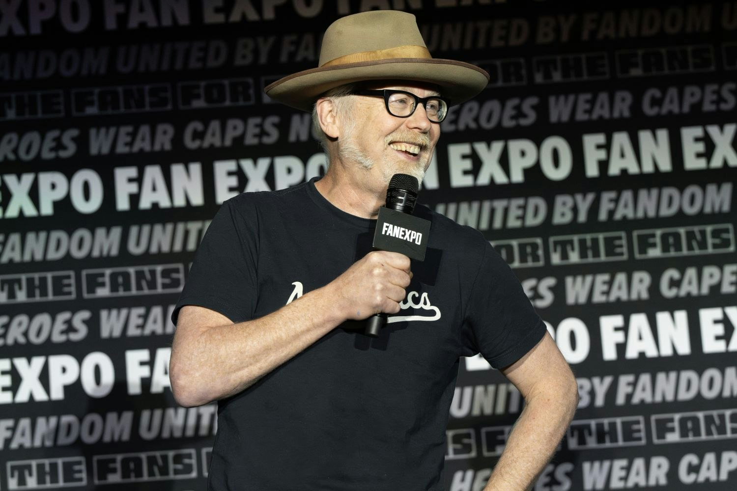 Adam Savage, with light skin, glasses, a trimmed white beard, and a tan wide-brimmed hat holds a microphone and smiles while speaking to the audience. He wears a black t-shirt with white script across the front. The FAN EXPO panel backdrop behind him includes phrases like “United by Fandom” and “Not All Heroes Wear Capes.”