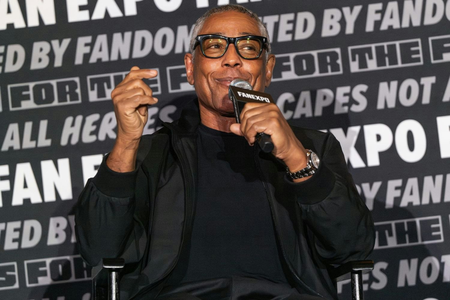 Giancarlo Esposito, with short gray hair sits in a director’s chair mid-sentence, smiling while holding a FAN EXPO mic. He wears black-rimmed glasses, a black jacket, black shirt, and a silver watch. His hand is raised slightly as he gestures expressively. The panel wall behind him features the FAN EXPO logo and fan slogans.