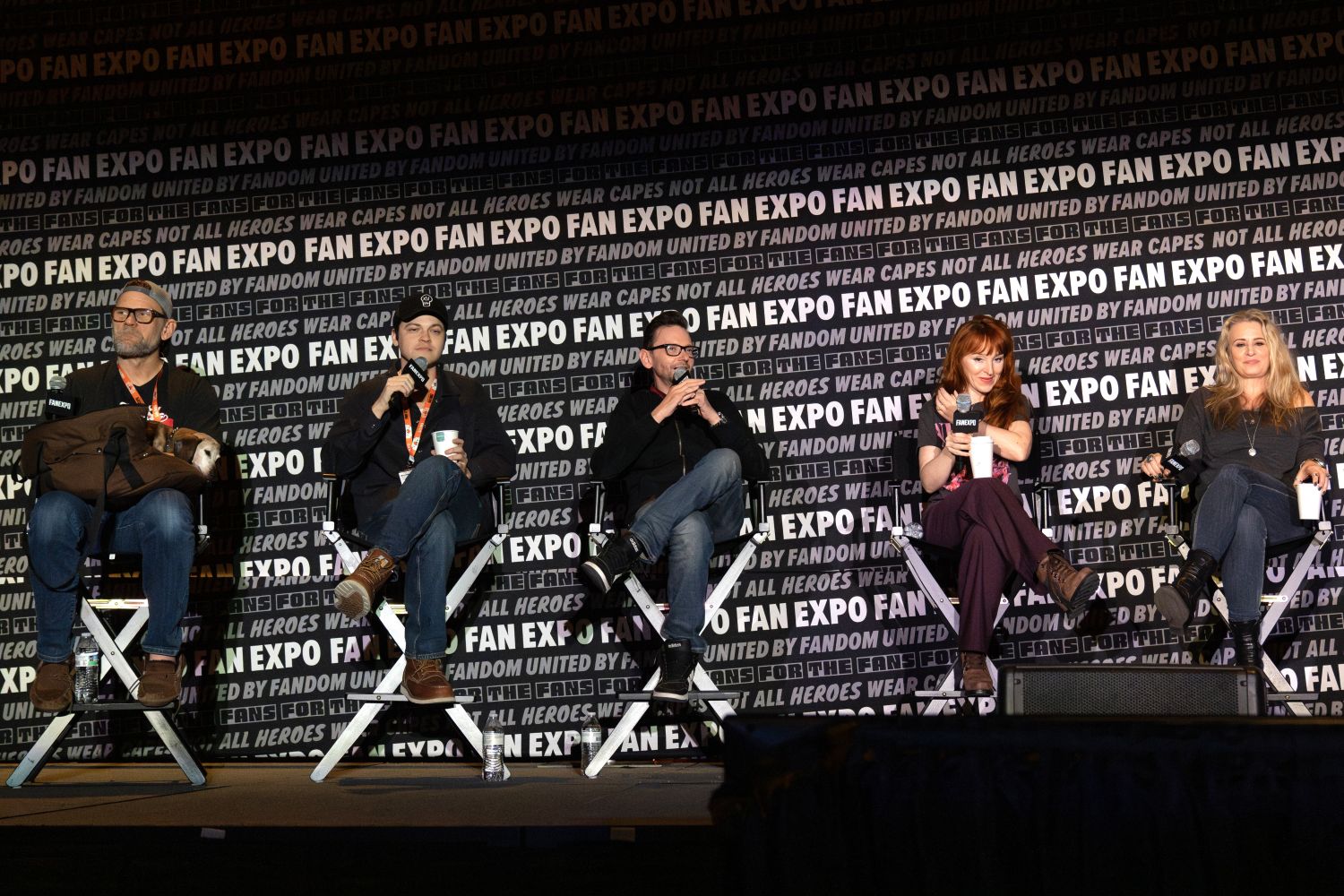 A panel of five Supernatural cast members sit onstage in director’s chairs during a group discussion. From left to right: Mark Pellegrino in a tan hat and glasses with a backpack in his lap, Alexander Calvert in a black hoodie and beanie sipping from a cup, DJ Qualls in dark glasses and black jacket speaking into a mic, Ruth Connell with long red hair wearing a pink and black top and burgundy pants, and Samantha Smith in a gray top holding a white mug. The stage is set against a bold black-and-white FAN EXPO backdrop.