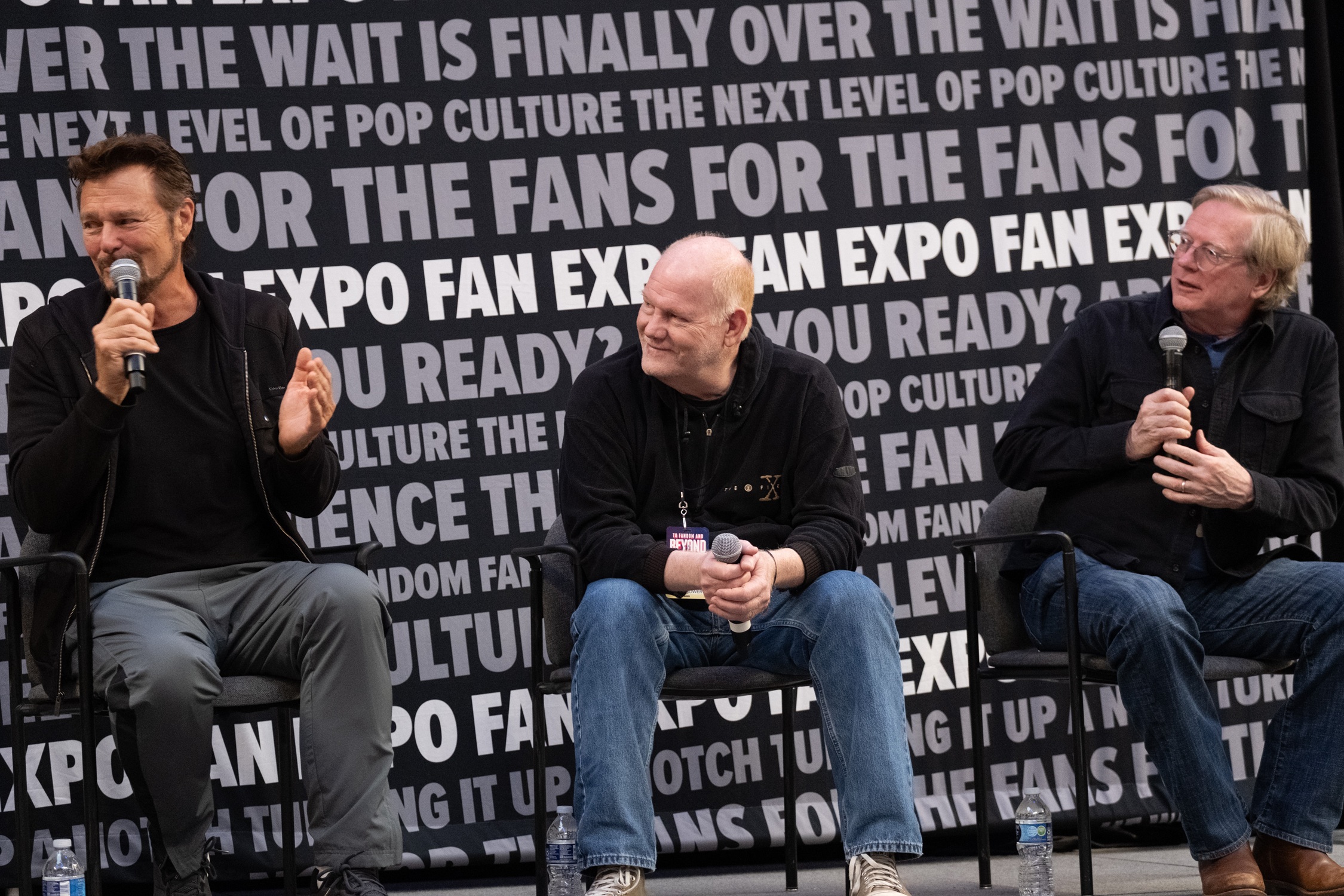 Greg Evigan, Glenn Morshower, and Dean Butler sit together on stage, microphones in hand, sharing behind-the-scenes stories and engaging with the audience.