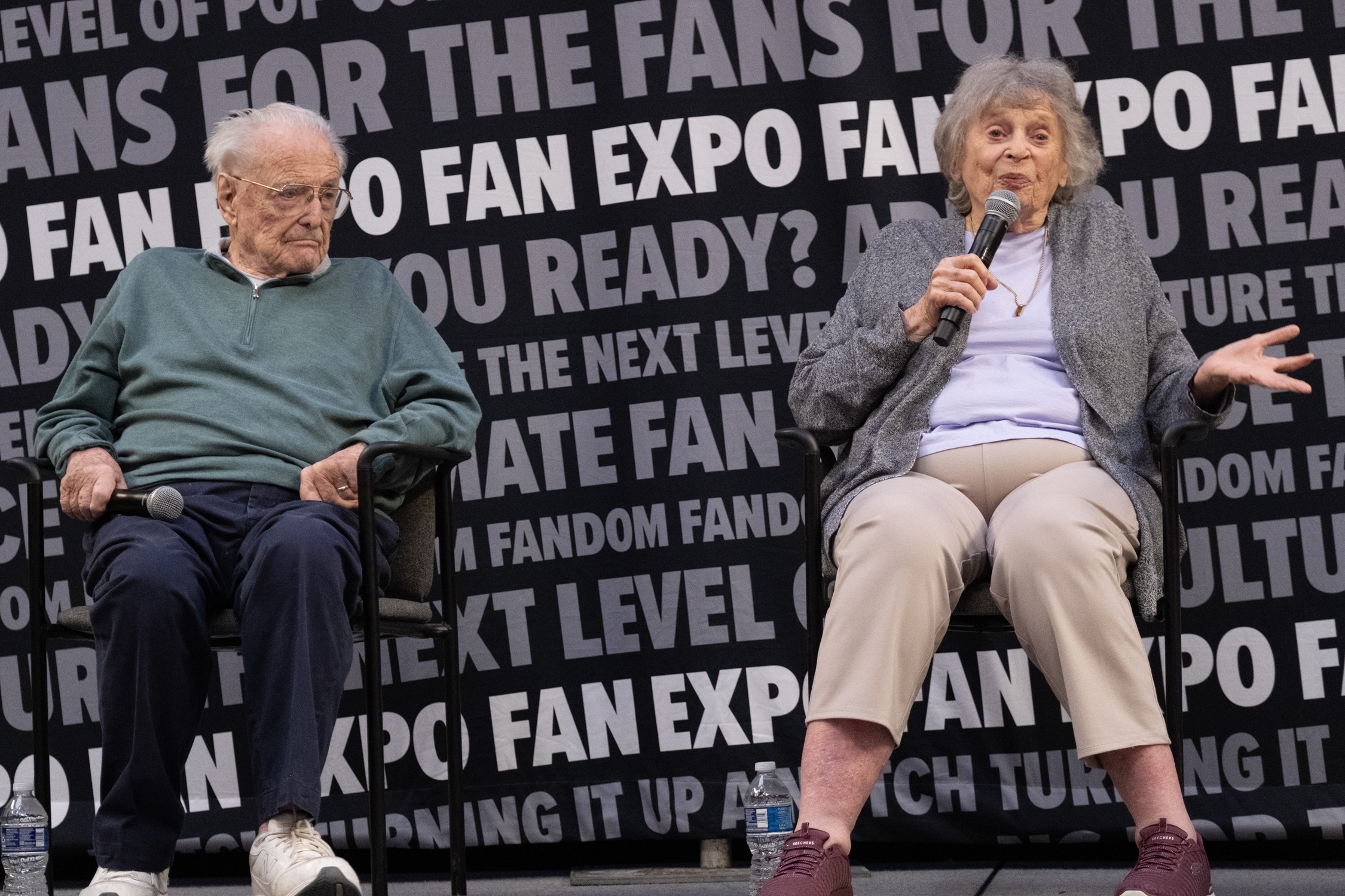 Actors William Daniels and Bonnie Bartlett sit side by side on stage during their panel at Dallas FAN FESTIVAL, microphones in hand as they share stories with the audience.