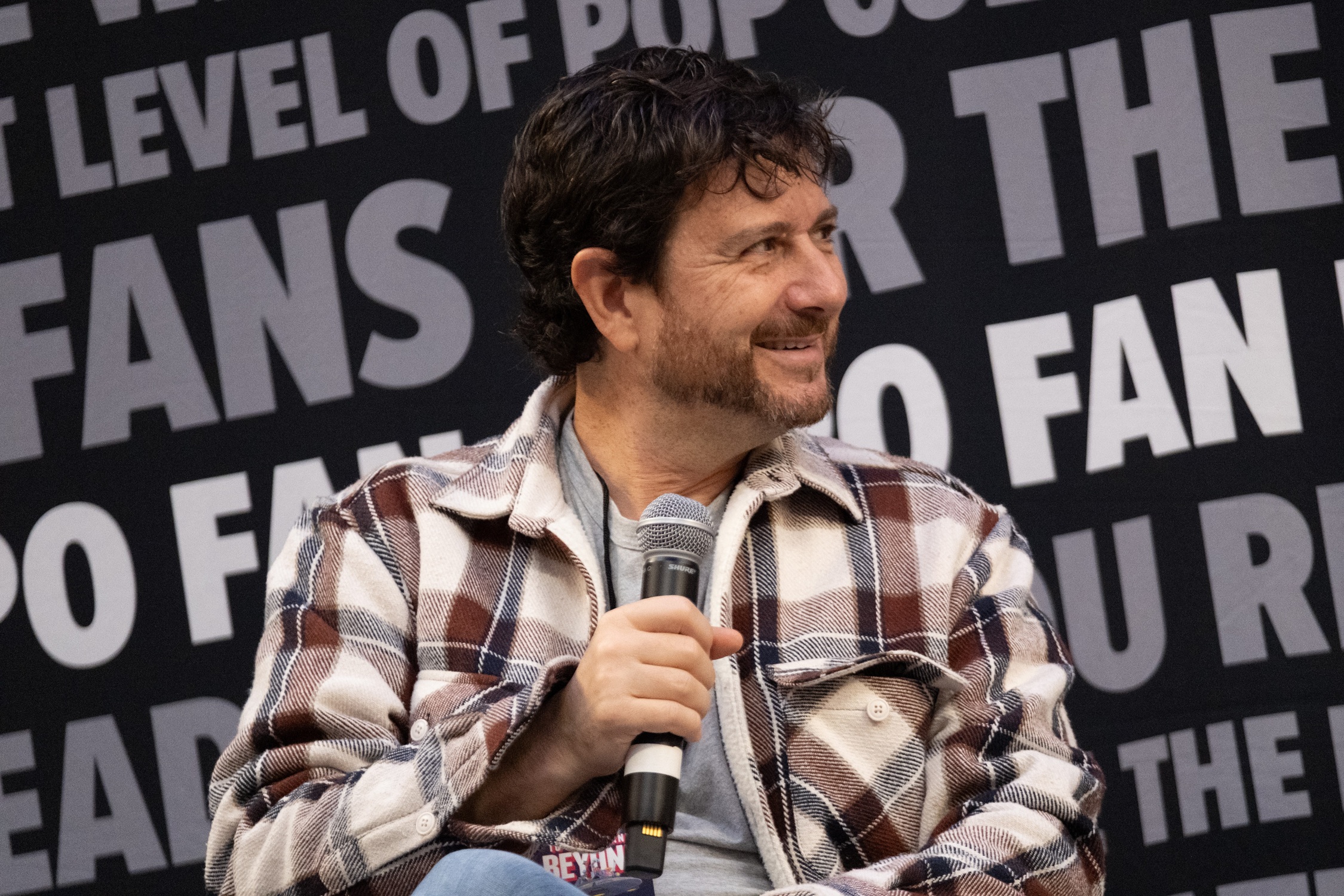Voice actor Rino Romano smiles on stage while speaking into a microphone during his spotlight panel, with the FAN EXPO backdrop behind him.