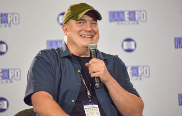 Brendan Fraser sitting on a stage, laughing into a microphone. He is wearing a navy fishing shirt with a black shirt underneath.