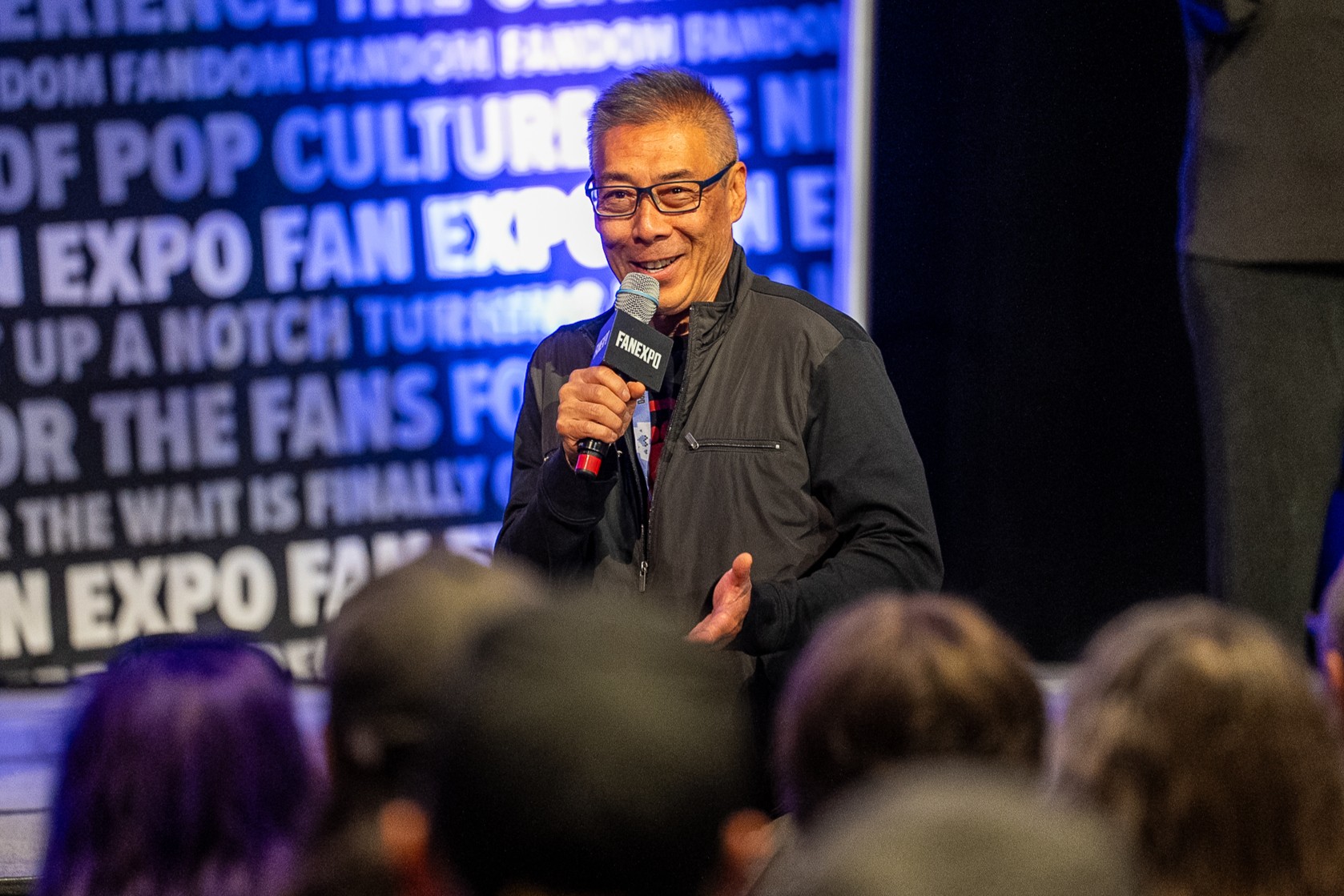 Francois Chau speaks on stage during a celebrity panel. Holding a microphone and smiling at the crowd, he shares stories while the audience listens intently.