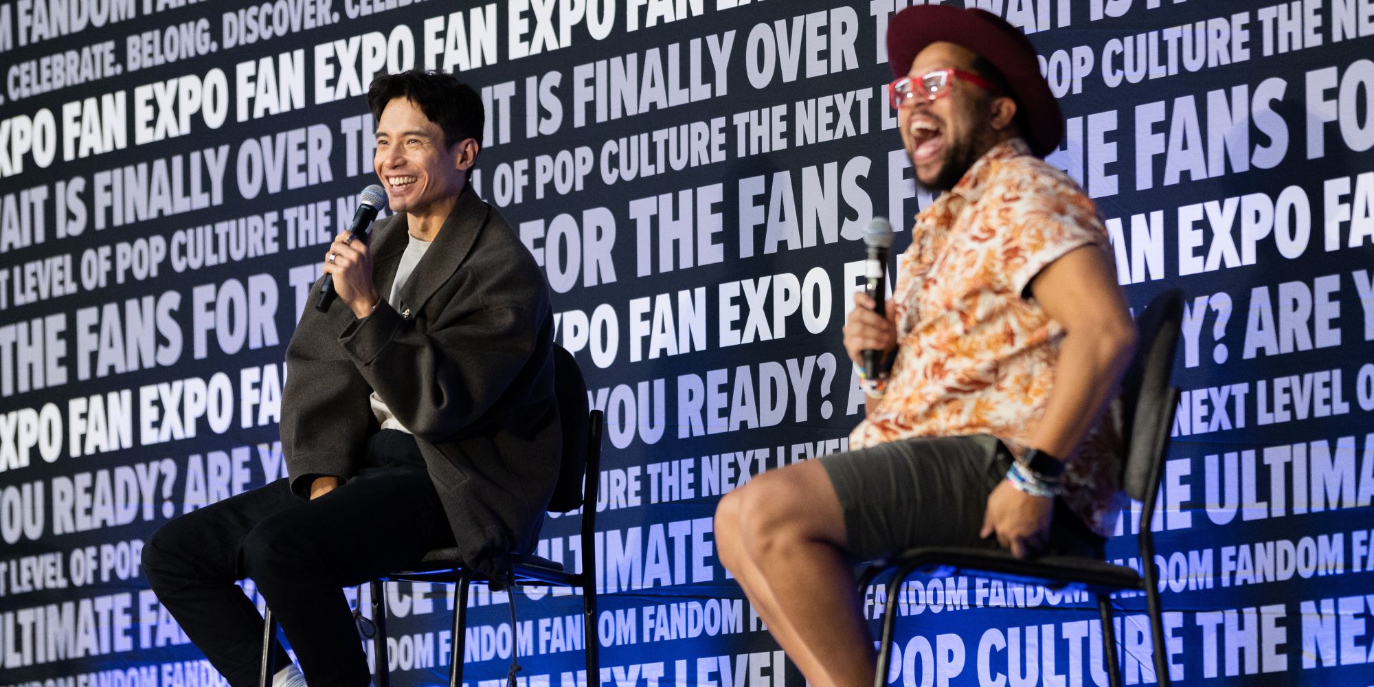 Two celebrity guests laugh and chat on stage during a Q&A session at Dallas FAN FESTIVAL, microphones in hand, as fans in the audience listen and cheer.