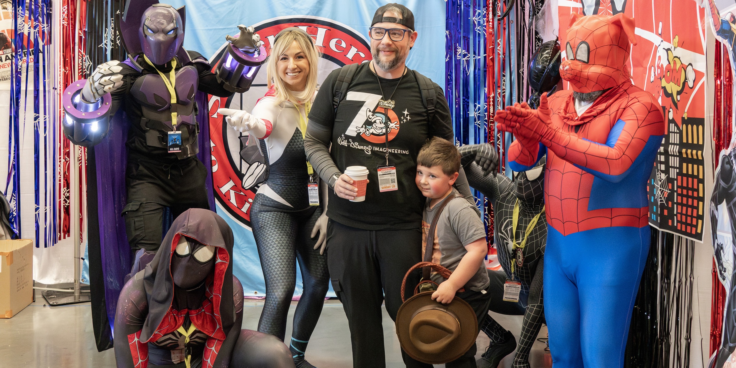 A group of adults and children pose together with costumed superheroes, including Spider-Man and armored characters. The group smiles while standing in front of colorful booth displays supporting charity and nonprofit organizations.