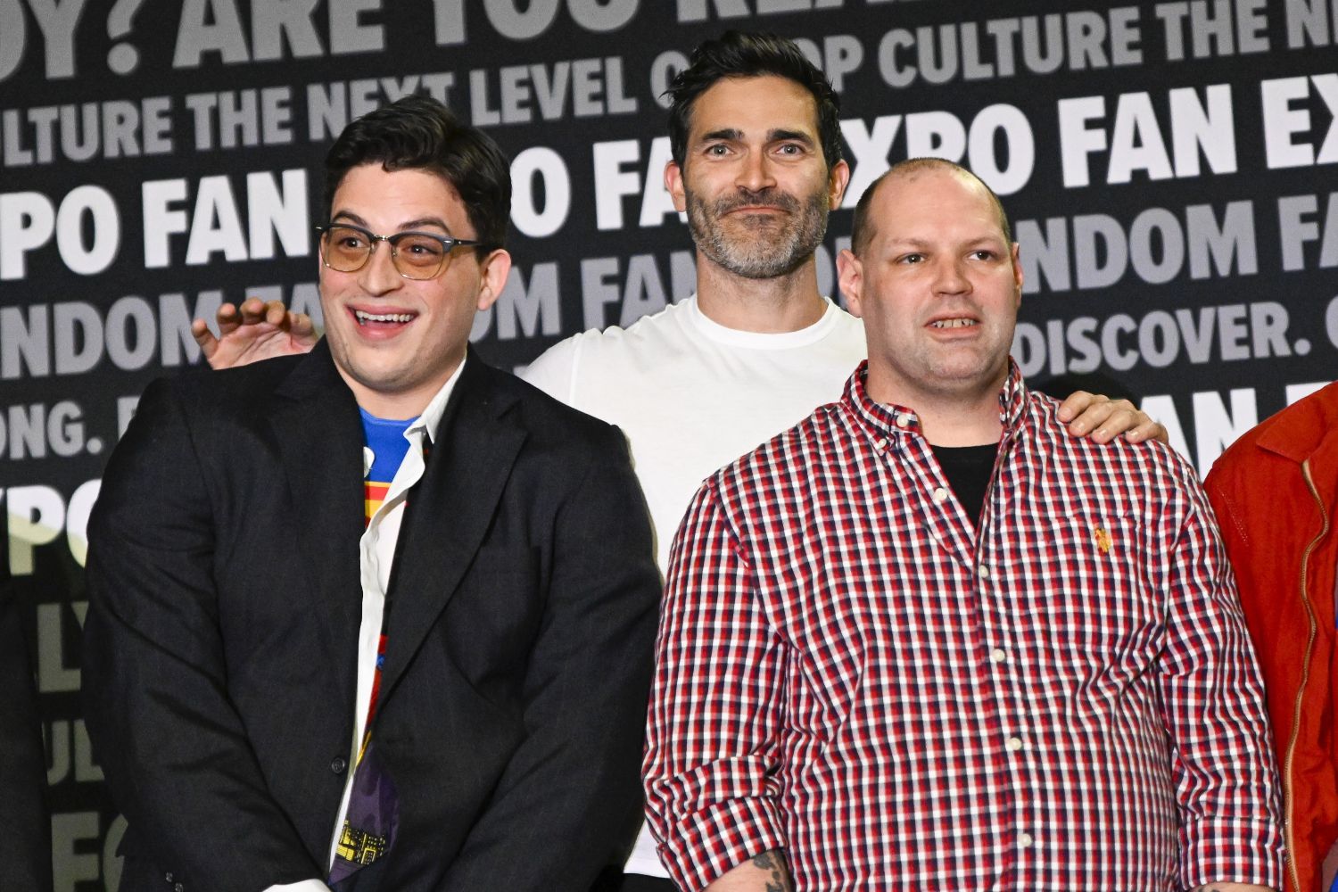 Three men pose for a candid group photo at the Clark Kent Look-Alike contest. The man on the left is in Clark Kent cosplay, smiling widely with his Superman shirt peeking out under a suit. In the center is actor Tyler Hoechlin, casually dressed in a white t-shirt. All three men stand against the FAN EXPO Cleveland backdrop.
