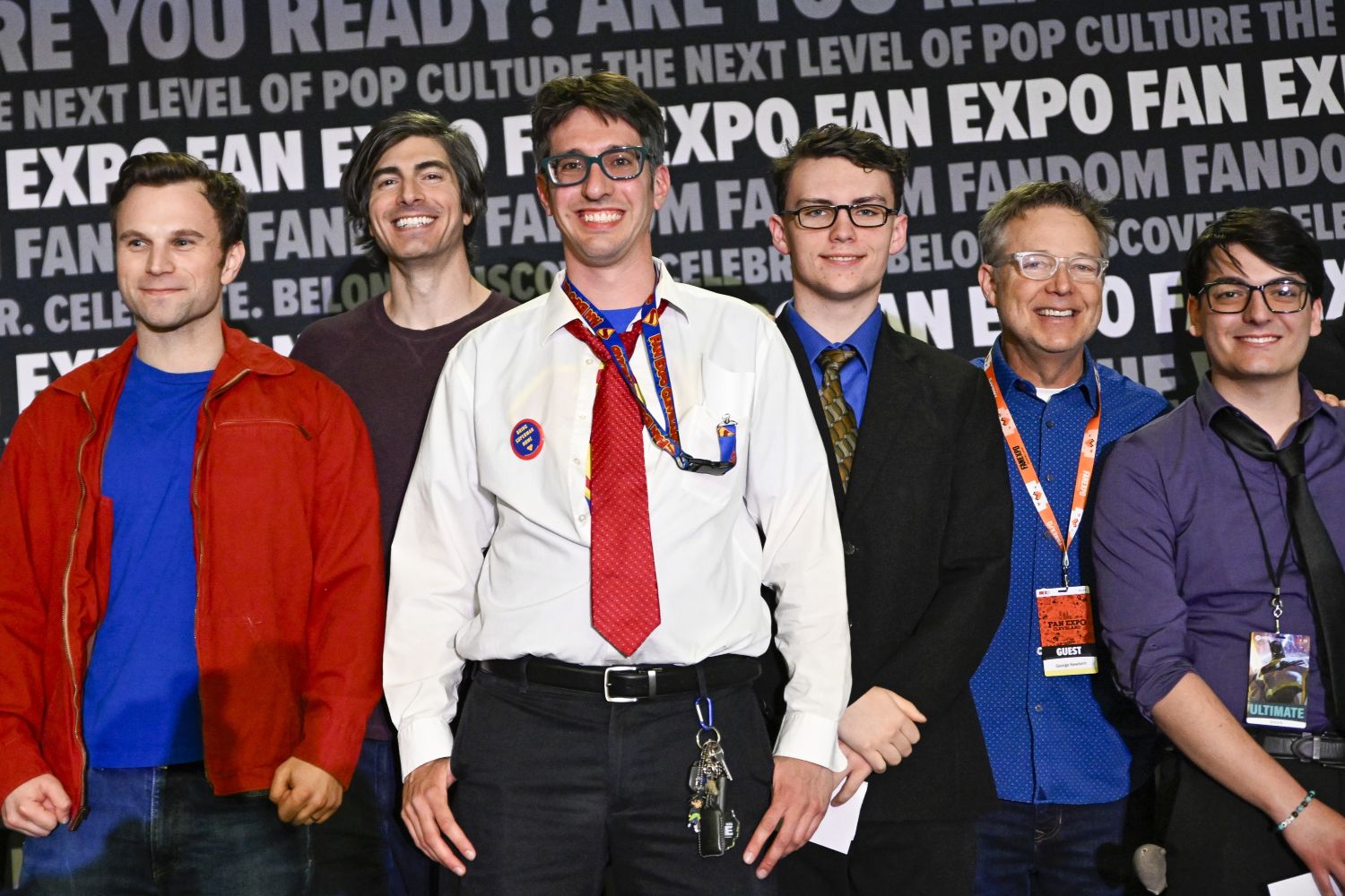 A group of six contestants lines up for a photo. The man in the center wears a loosely buttoned shirt that reveals a Superman logo tee beneath, complete with a red tie and Clark Kent glasses. He's flanked by fans and guests, including Brandon Routh (second from the left) and George Newbern (second from the right), all smiling proudly before the look-alike contest.
