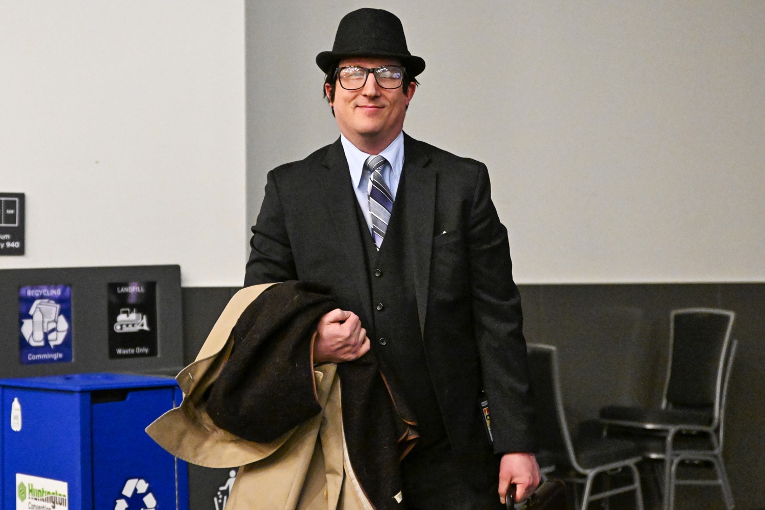 A man stands indoors wearing a black three-piece suit, striped tie, and bowler hat. He’s holding a trench coat and briefcase, emulating a 1950s Clark Kent look. He’s smiling softly at the camera, posed near a recycling station in a hallway outside the main event space.