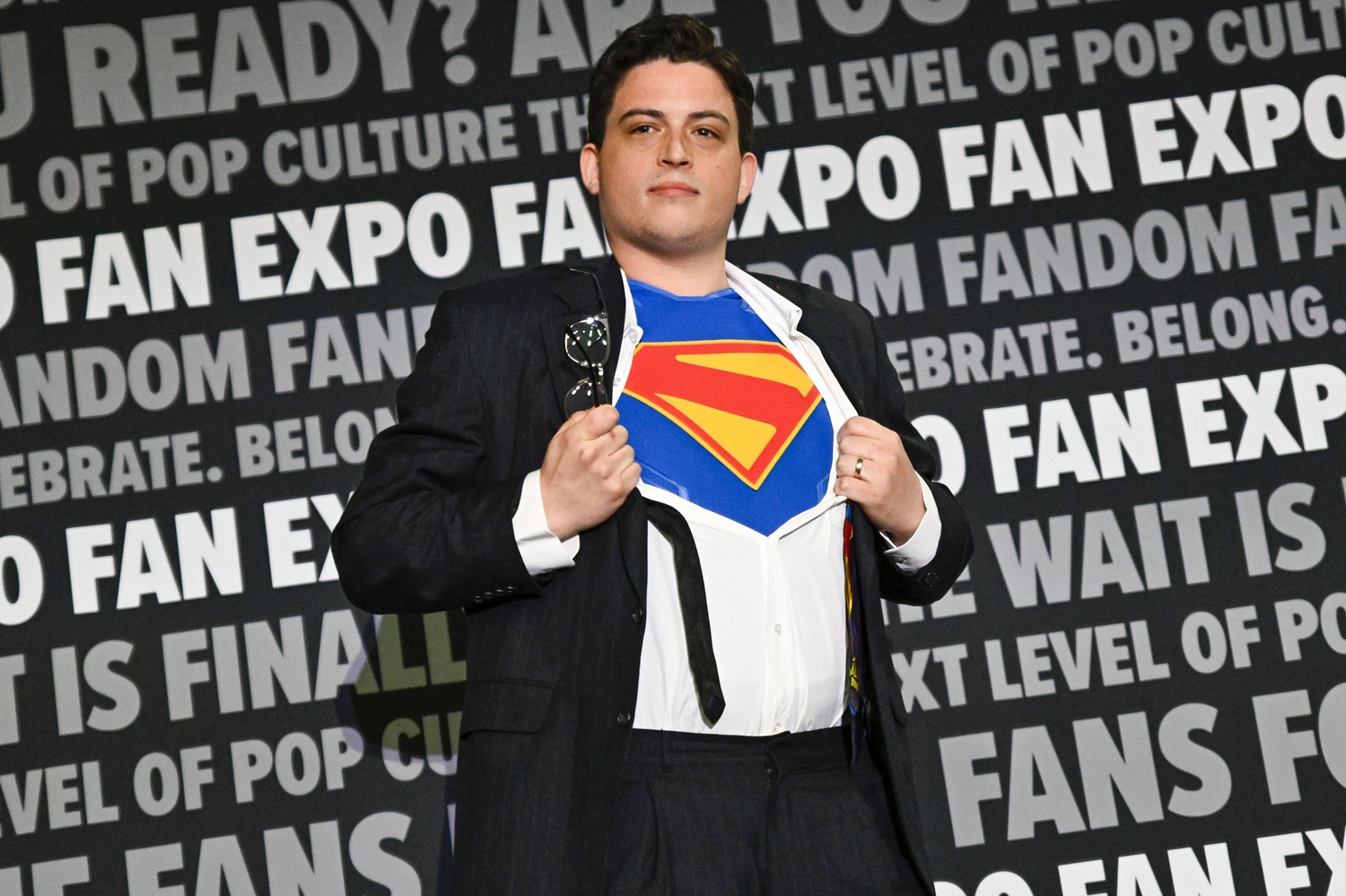 A man on stage tugs open his button-down shirt to reveal a bright Superman logo underneath. He wears a black suit and holds glasses in his right hand, evoking the classic comic book transformation from Clark Kent to Superman. The FAN EXPO step-and-repeat backdrop is behind him, covered in bold text about fandom and pop culture.