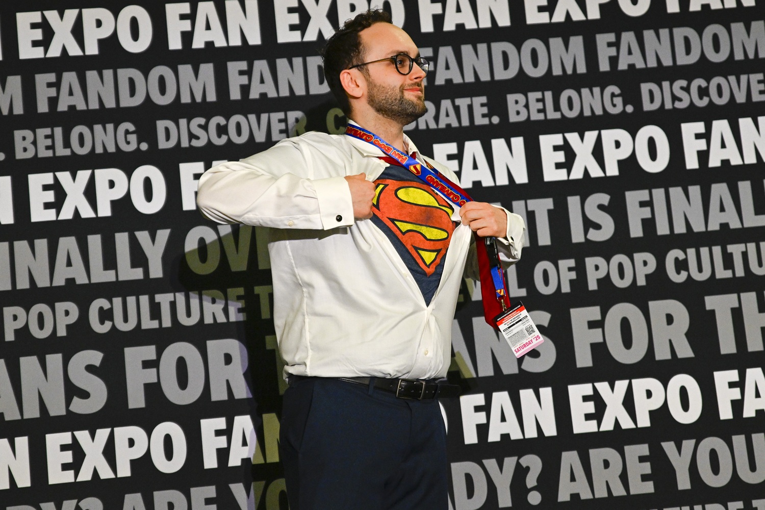 A smiling man wears dress pants and a white button-down shirt, which he dramatically opens to reveal a Superman t-shirt underneath. His FAN EXPO badge and Superman-themed lanyard swing as he strikes a heroic pose against the branded backdrop.