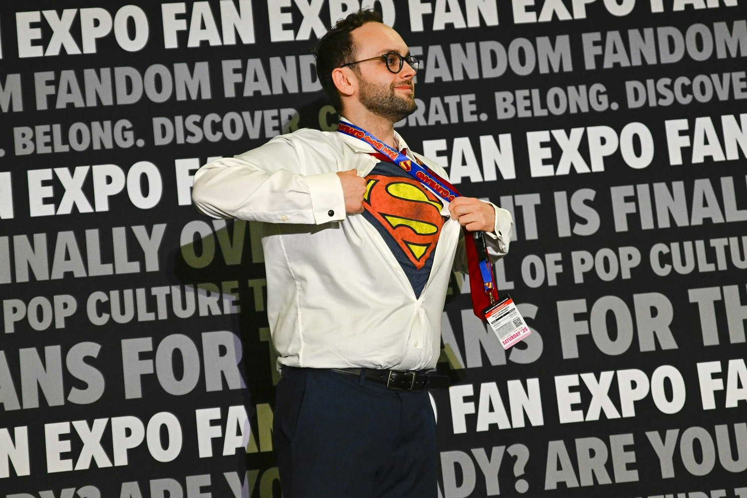 A smiling man wears dress pants and a white button-down shirt, which he dramatically opens to reveal a Superman t-shirt underneath. His FAN EXPO badge and Superman-themed lanyard swing as he strikes a heroic pose against the branded backdrop.