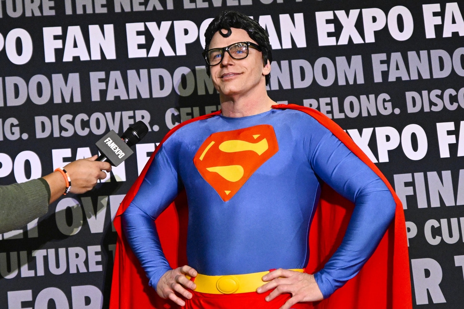A cosplayer fully suited as Superman — blue bodysuit with a red cape, yellow belt, and signature “S” emblem — poses with hands on hips and a confident grin. He wears a black curl wig and square glasses in homage to Clark Kent. A FAN EXPO microphone is being held up to him for an interview.