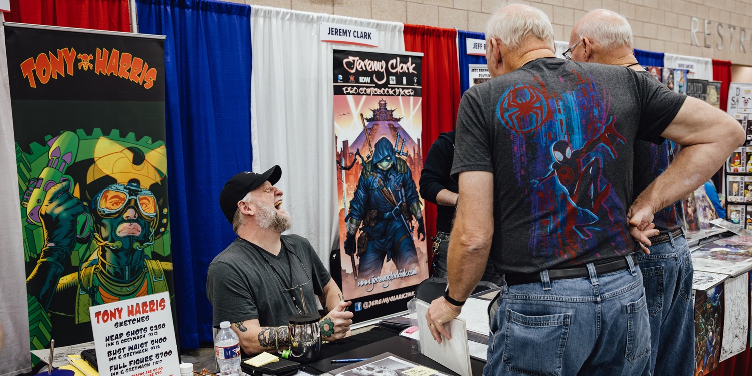 Comic book artist Tony Harris laughs while speaking with fans at his booth in Artist Alley at Dallas FAN FESTIVAL. Behind him, colorful banners featuring comic art and character illustrations are displayed.